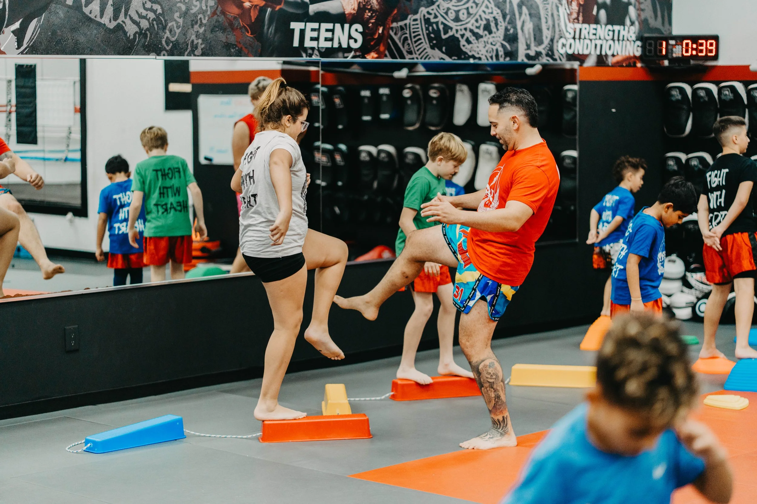 An instructor assists children in a martial arts or fitness class, guiding them through a balance or coordination exercise using small platforms in a gym with boxing gloves and equipment on the wall.