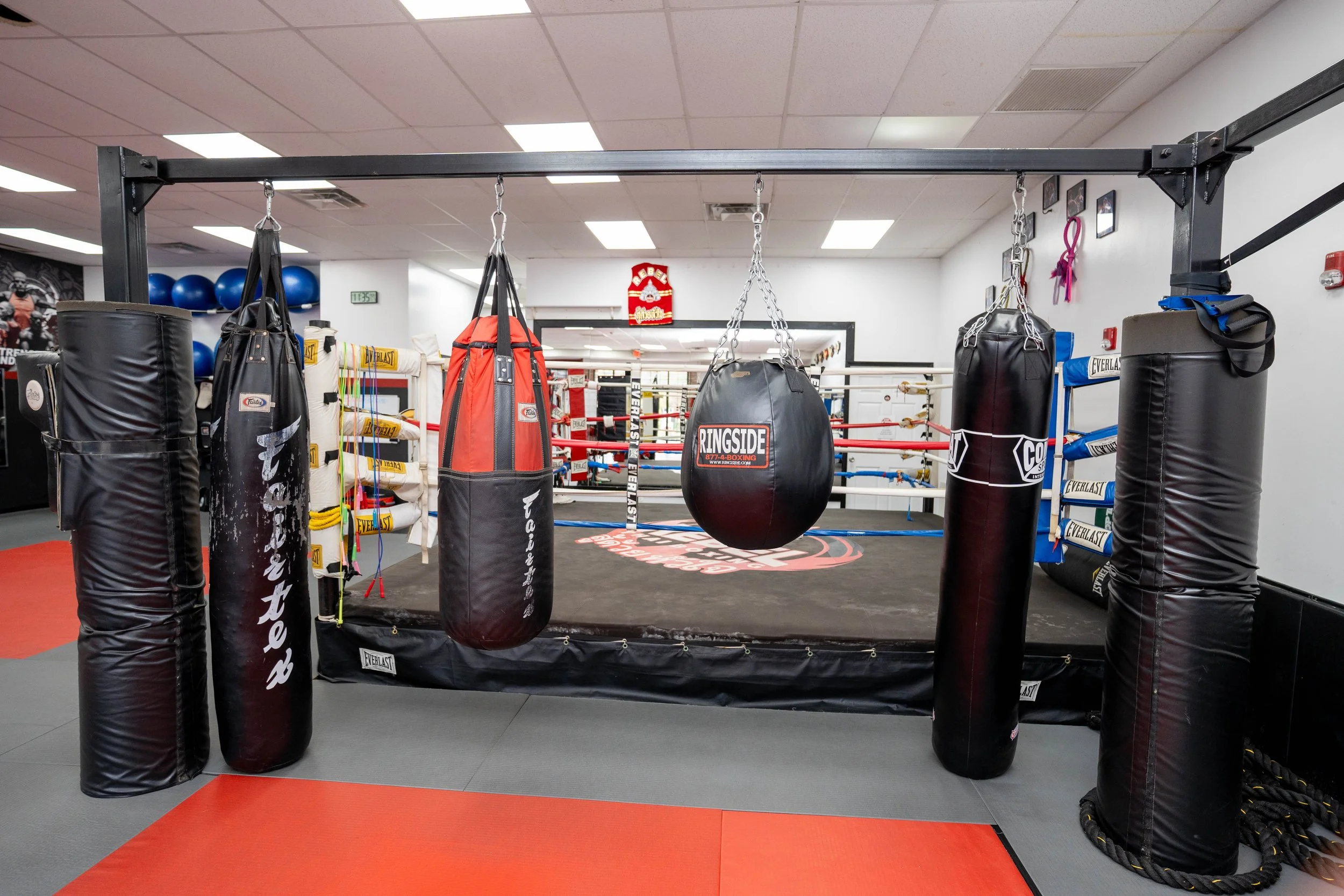 A boxing gym with a boxing ring surrounded by punching bags, including speed bags, hanging inside the ring. The gym has padded flooring with red and gray sections, and various gym equipment like medicine balls and resistance bands are visible.
