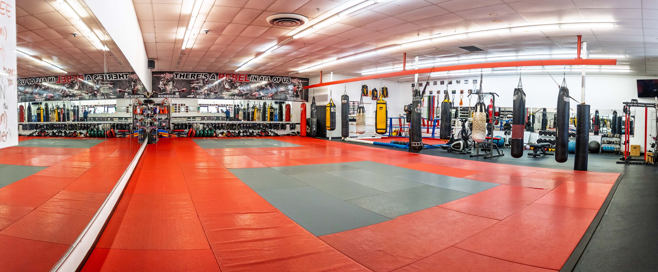 Empty boxing gym with red and gray mats, punching bags, and workout equipment stored on shelves.