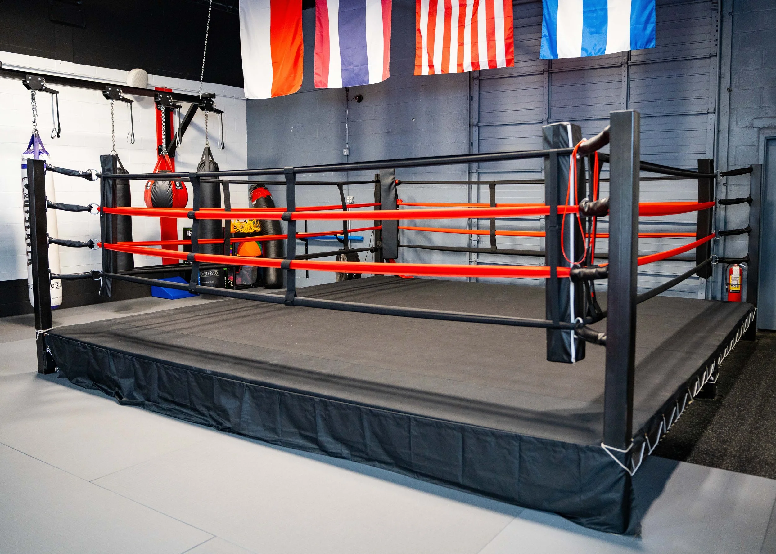An empty boxing ring inside a gym with flags hanging above, black walls, and various punching bags and equipment in the background.