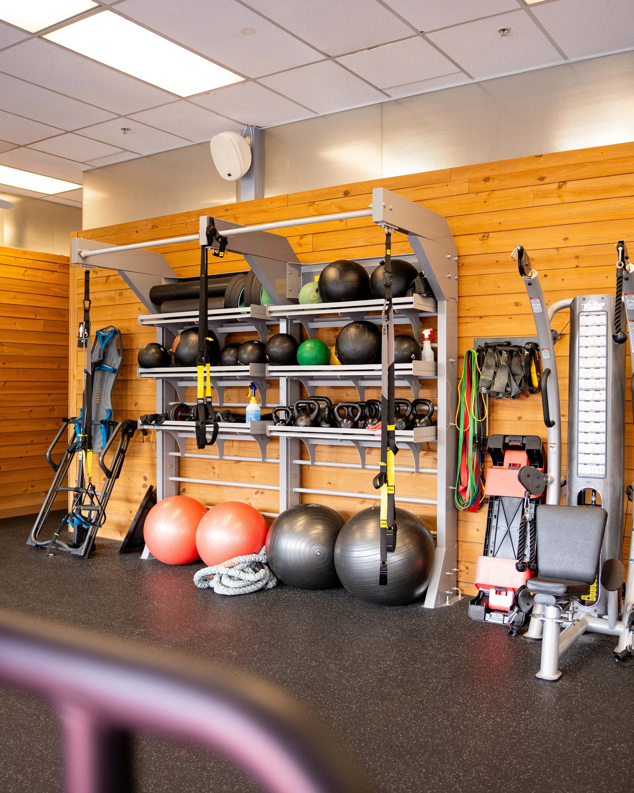 Organized gym equipment on shelves and the floor, including exercise balls, kettlebells, resistance bands, a spray bottle, and various workout accessories, with a wooden wall background.