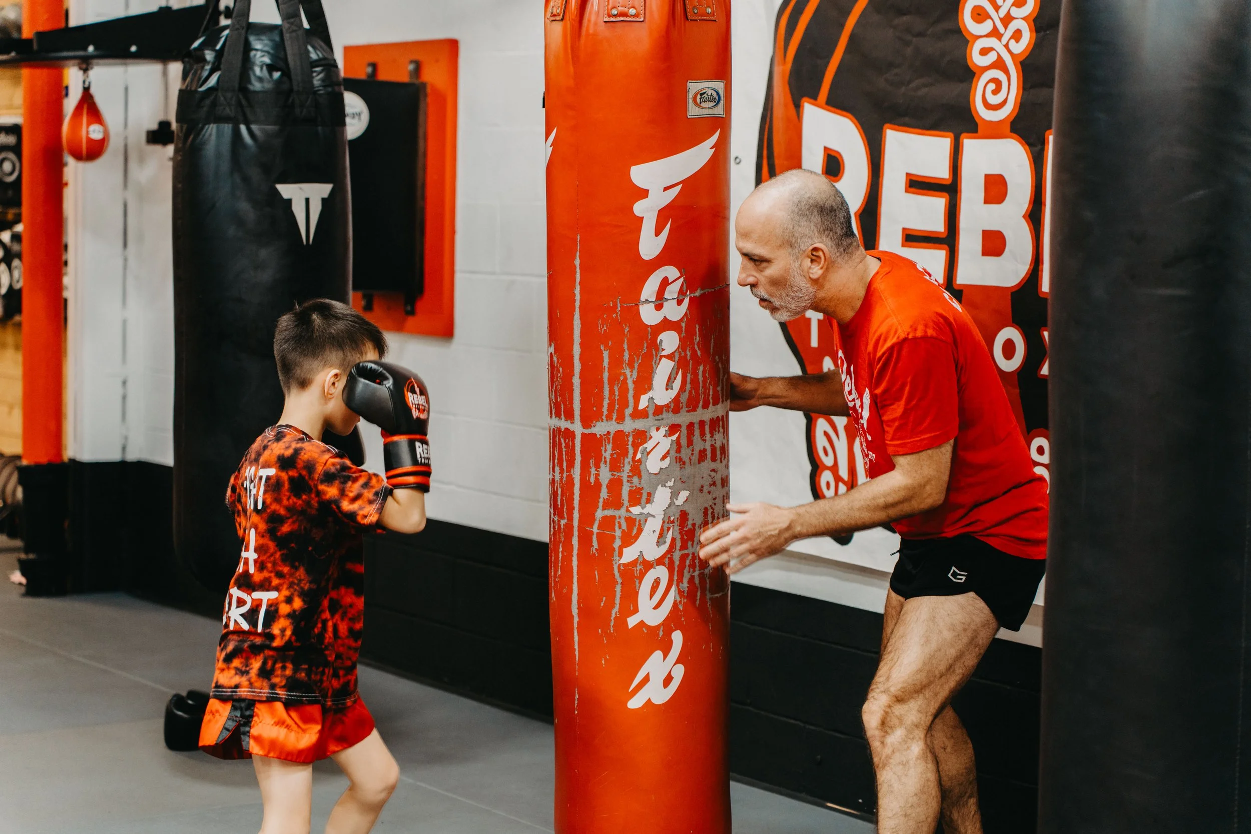 A young boy practicing boxing with a coach in a gym, wearing boxing gloves and a tie-dye t-shirt with red shorts, while an older male trainer is demonstrating techniques near a red punching bag.