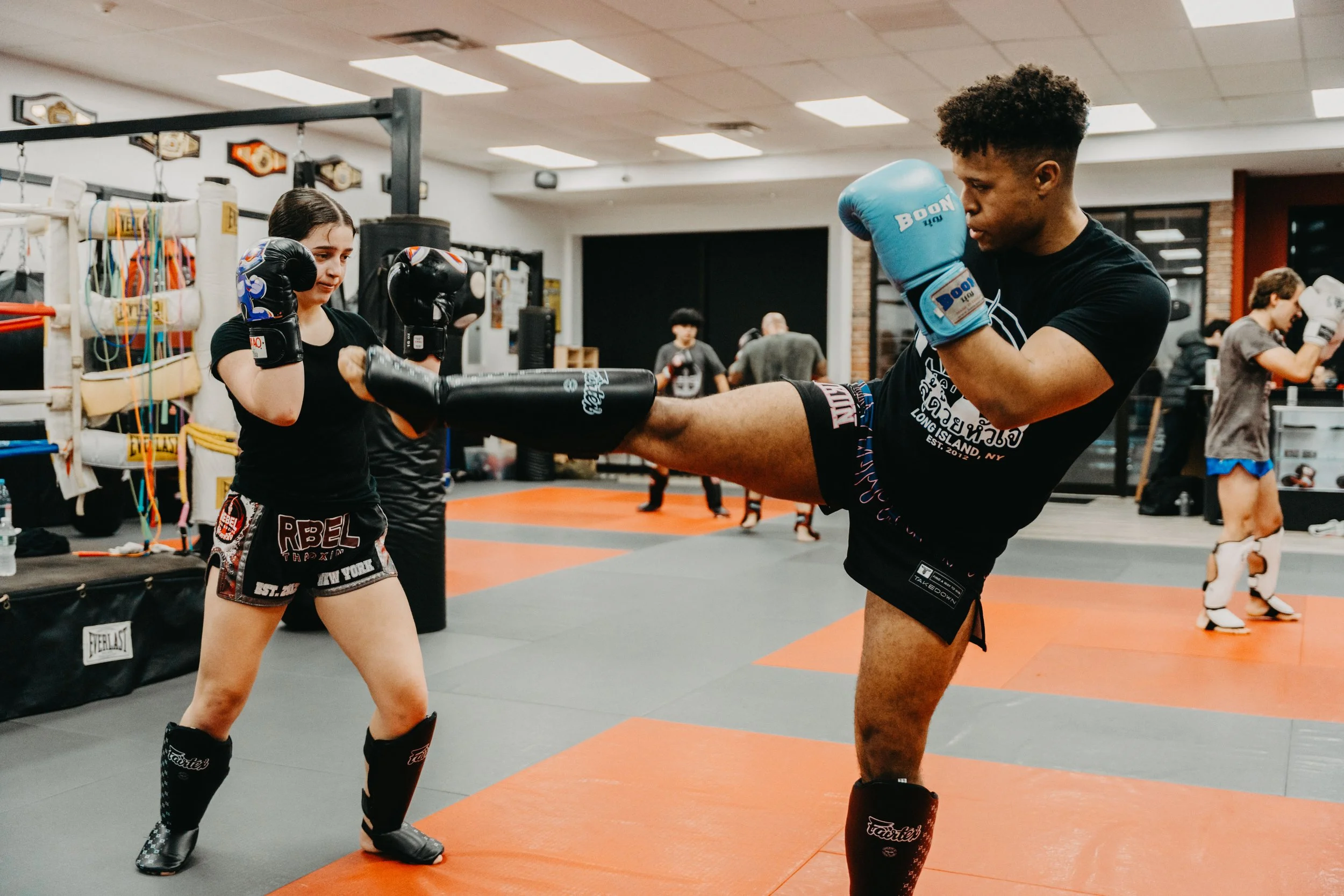 A male martial artist practicing a high kick with his right leg aimed at a female opponent's midsection in a gym. The female is holding focus mitts and wearing black shorts and gloves, while the male is wearing blue gloves, black shorts, and shin gua