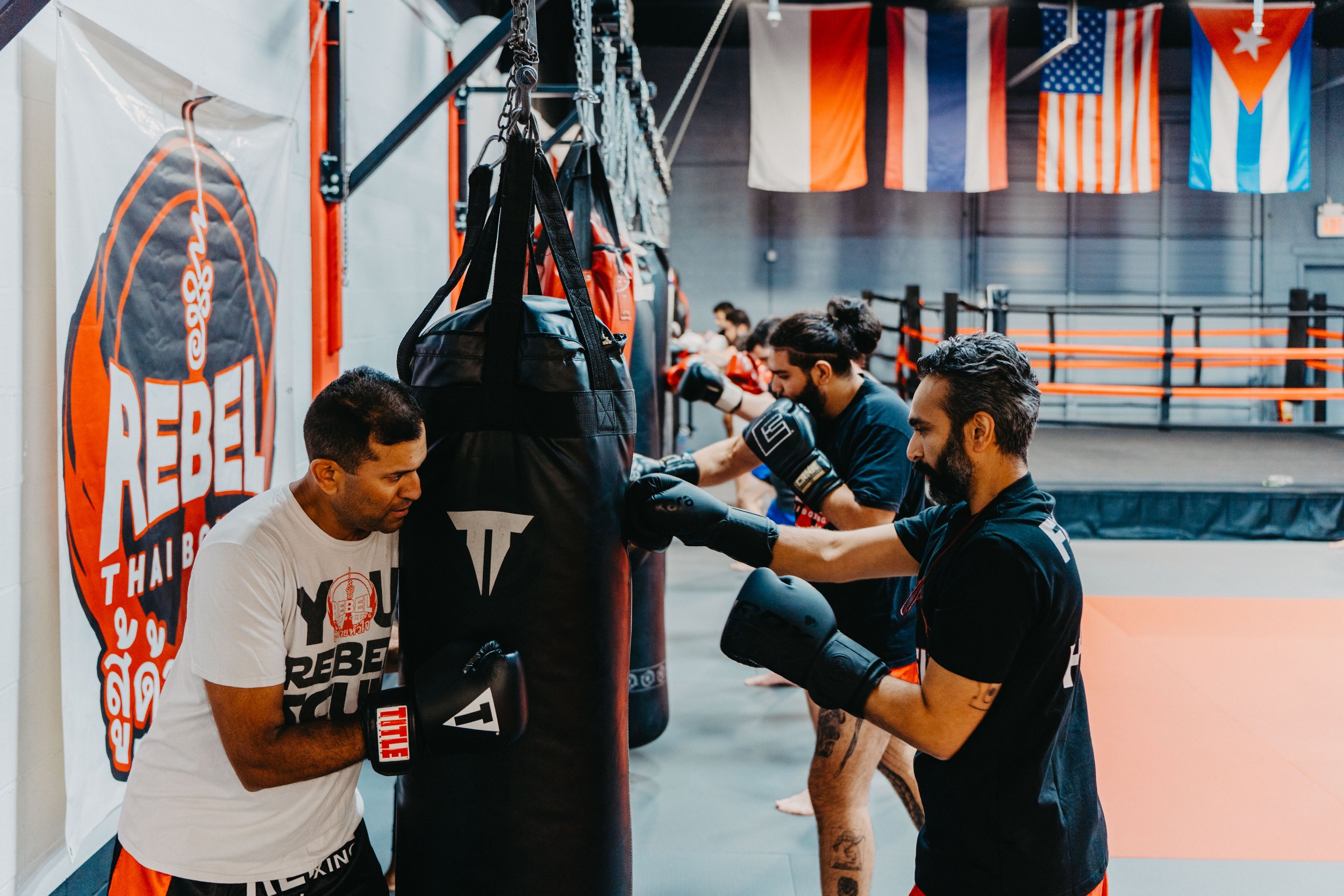Group of men practicing boxing in a gym, some throwing punches at a hanging punching bag, with flags hanging from the ceiling.