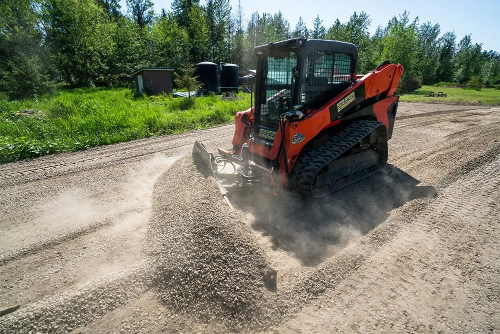 A skid steer loader moving gravel on a dirt road in a rural area with green trees and a small building in the background.