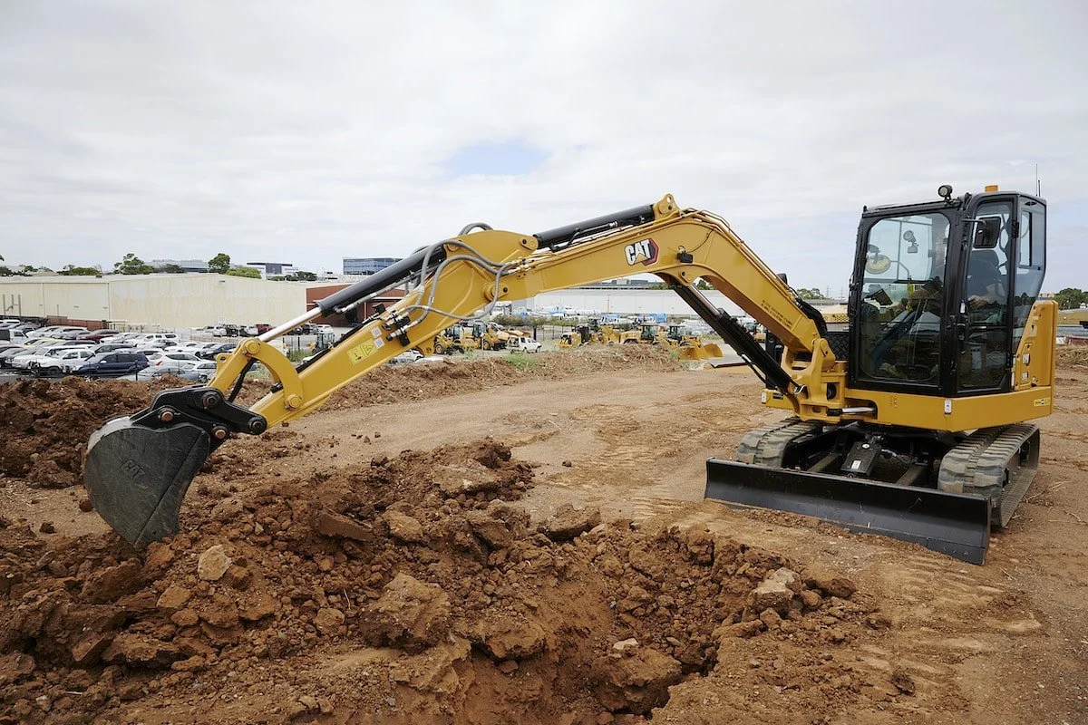 A yellow Caterpillar excavator digging into a mound of dirt at a construction site with a parking lot and industrial buildings in the background.