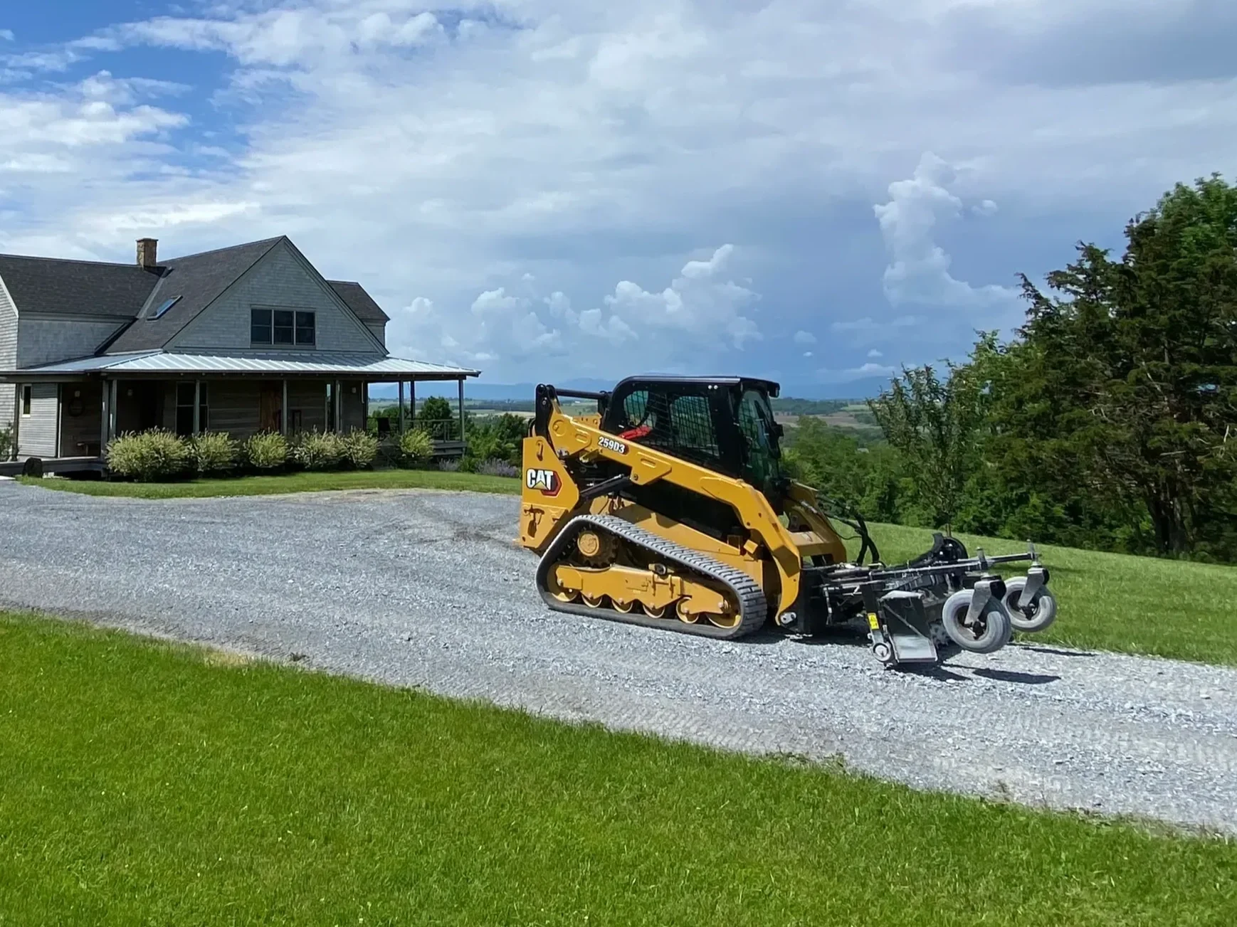 A yellow Caterpillar skid-steer loader on a gravel driveway in front of a gray house, with green grass and trees, under a partly cloudy sky.
