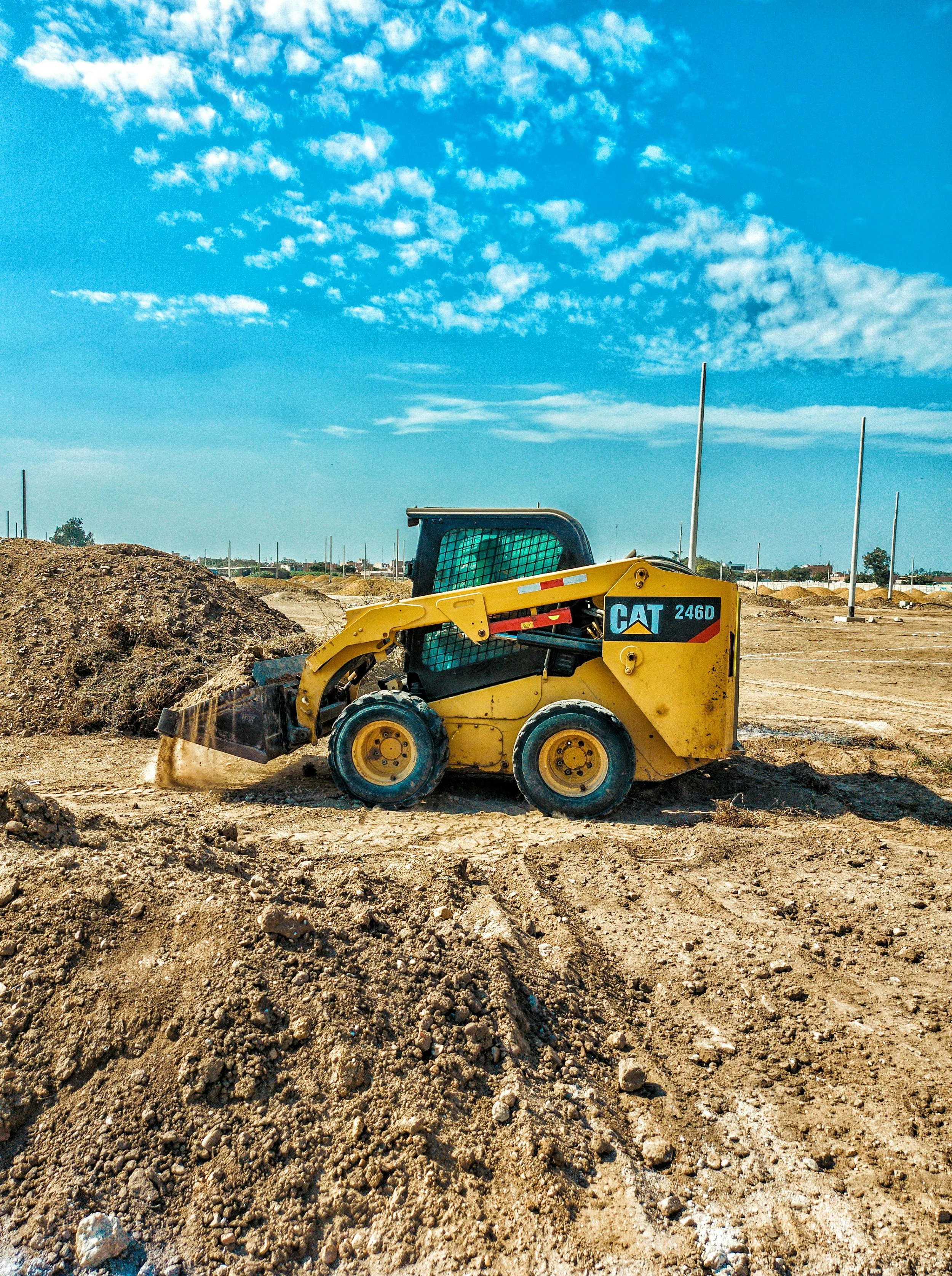 A yellow CAT 246D skid steer loader moving dirt on a construction site with a cloudy blue sky in the background.
