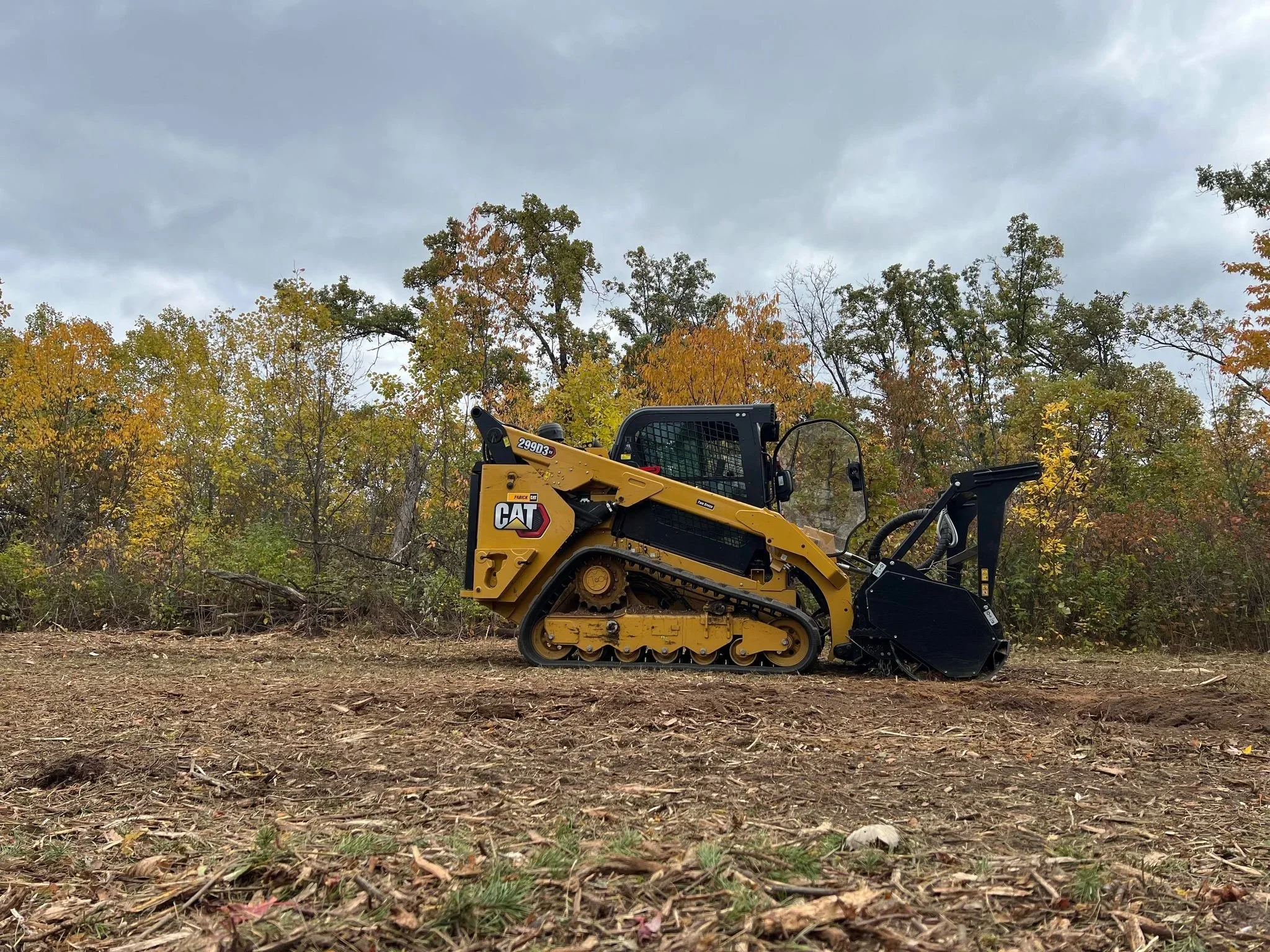 A yellow and black Caterpillar track loader on dirt ground, with a background of trees with autumn foliage under a cloudy sky.