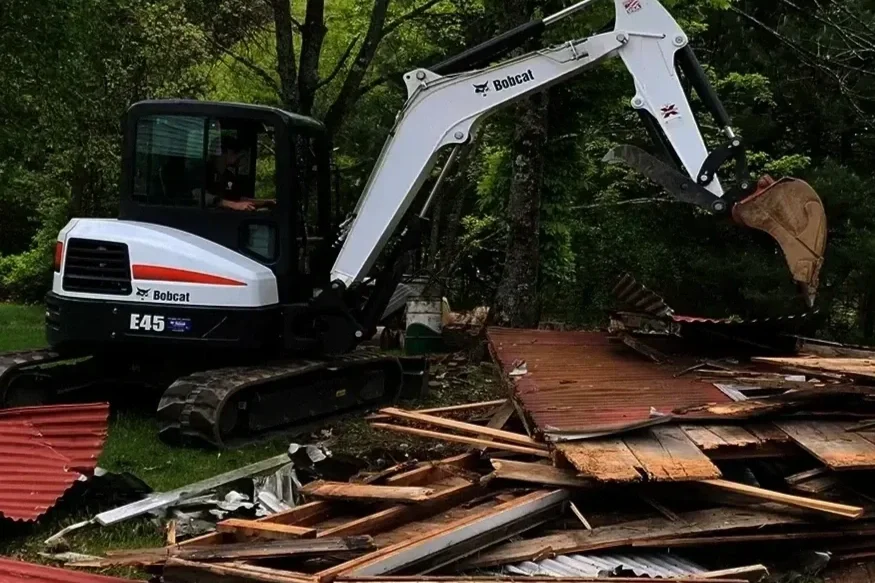 A small white Bobcat E45 mini excavator with black tracks is demolishing a wooden structure with a bucket attachment, surrounded by trees and debris.