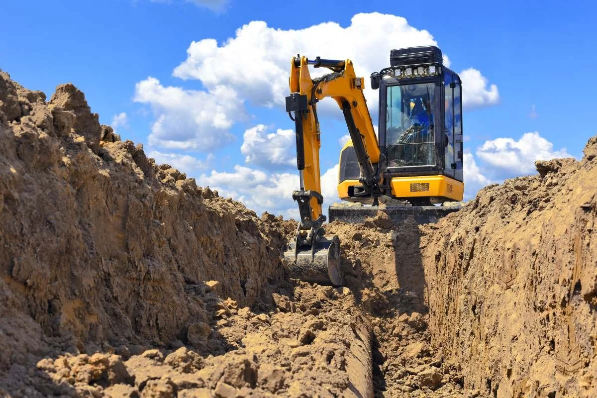 A yellow excavator working in a deep trench on a construction site under a blue sky with clouds.