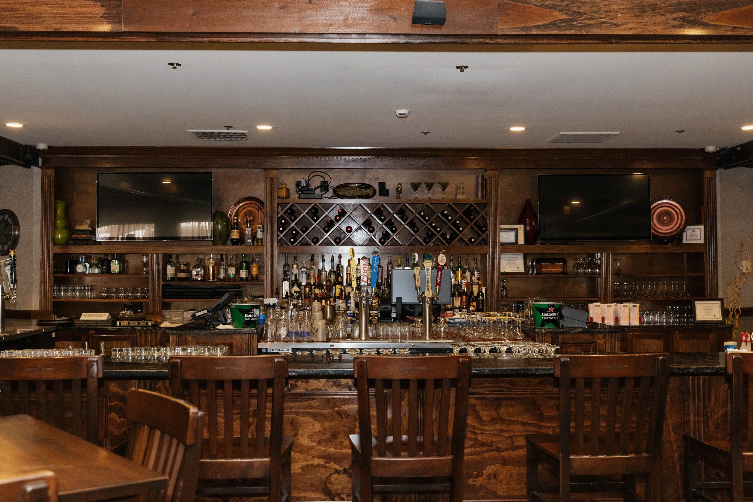 Dark wooden bar with shelves stocked with various bottles of alcohol, two television screens, and bar equipment. Wooden chairs are in front of the bar, and the background includes wine racks and decorative items.