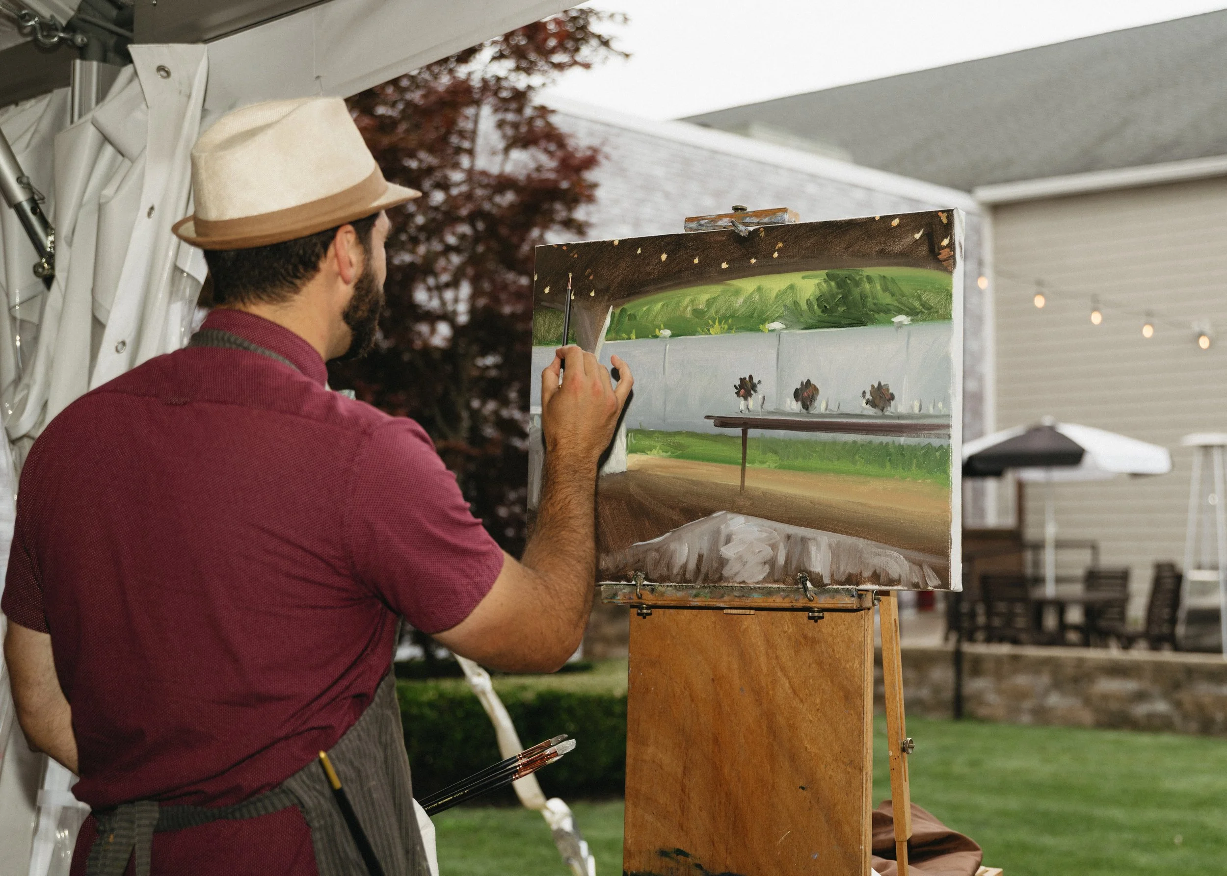 A man with a fedora hat painting a landscape on an easel outdoors. The landscape includes trees, a field, and a cloudy sky.