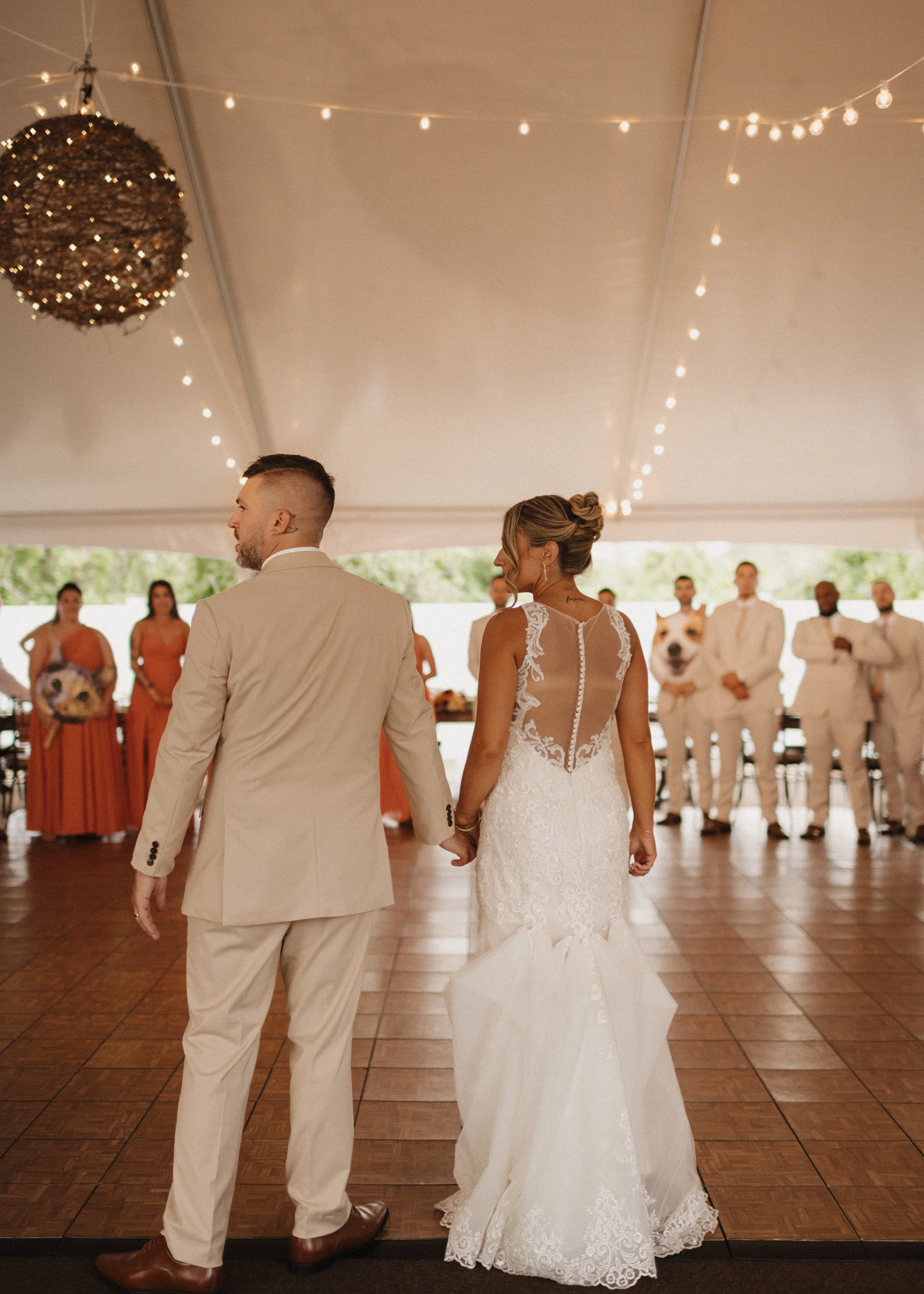Bride and groom holding hands during their wedding reception under a decorated tent with string lights and a chandelier, with guests in the background.