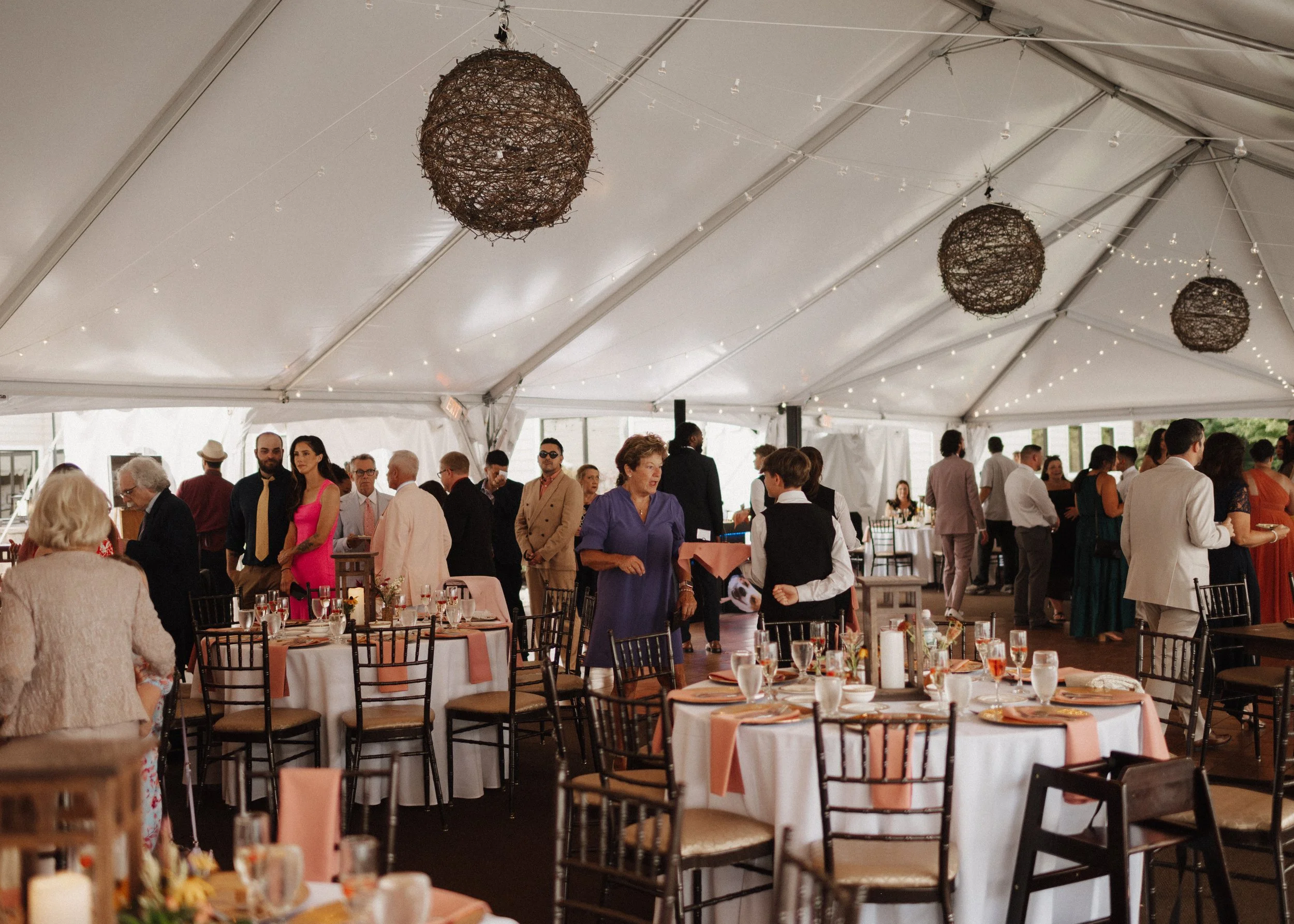 Guests at a formal gathering under a white tent, with decorated tables, hanging lights, and people socializing.