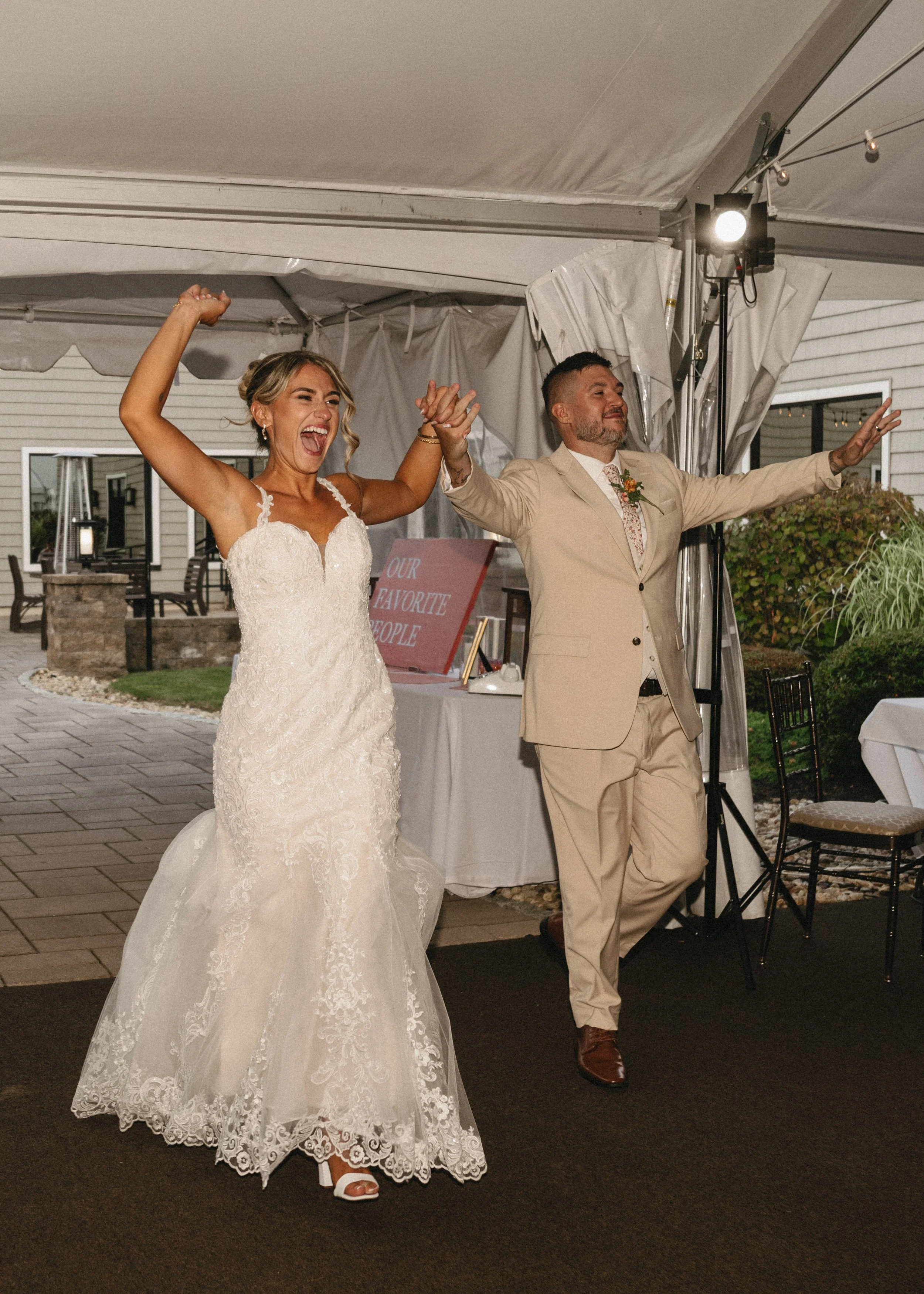 Bride and groom celebrating at their wedding reception under a tent, with the bride in a white lace wedding gown and the groom in a beige suit, holding hands and smiling joyfully.