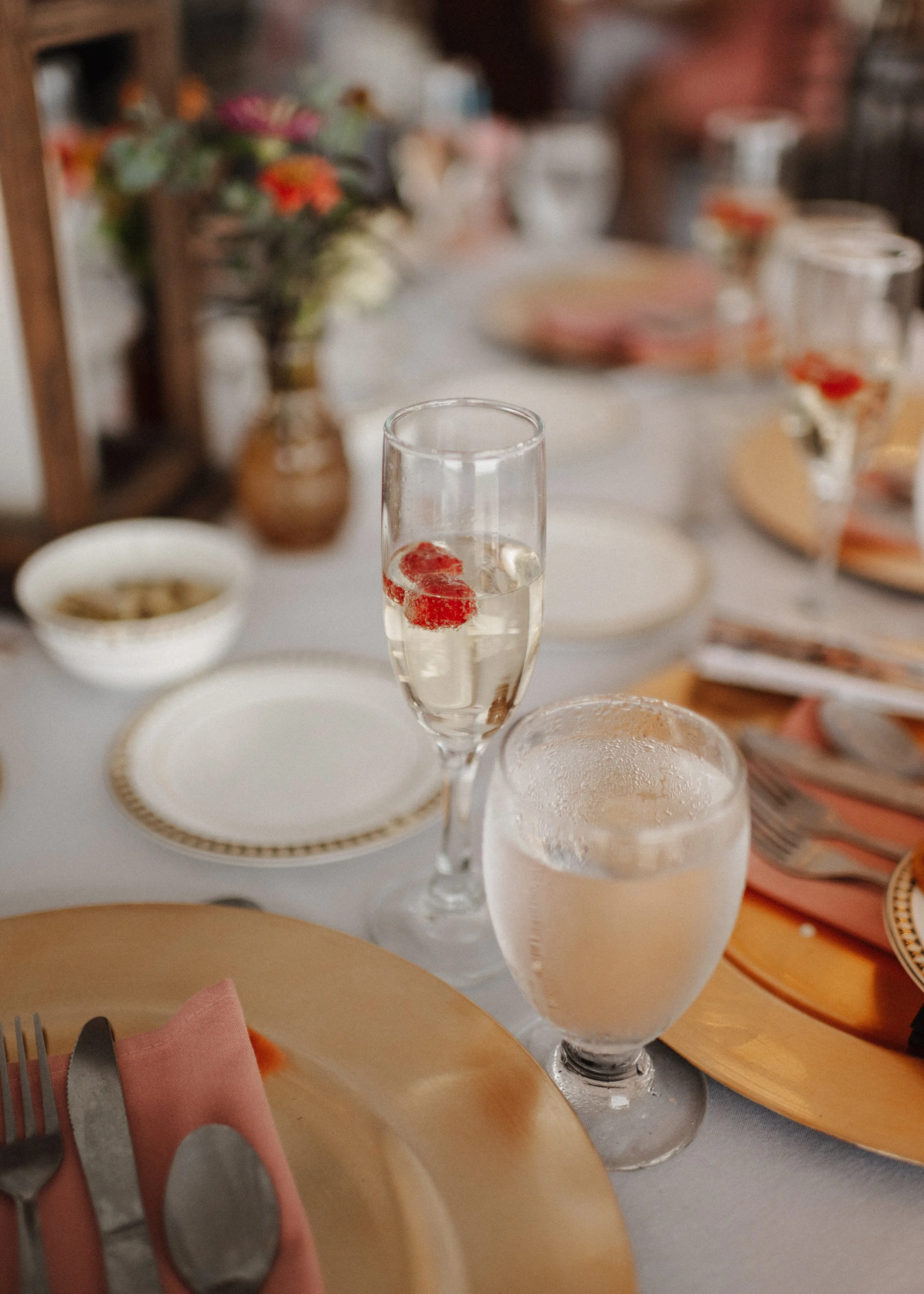 A close-up of a table setting with a champagne flute filled with clear liquid and a few strawberries, a glass of water, a pink napkin with cutlery, and a striped plate on a white tablecloth. In the background, there is a blurred flower arrangement an