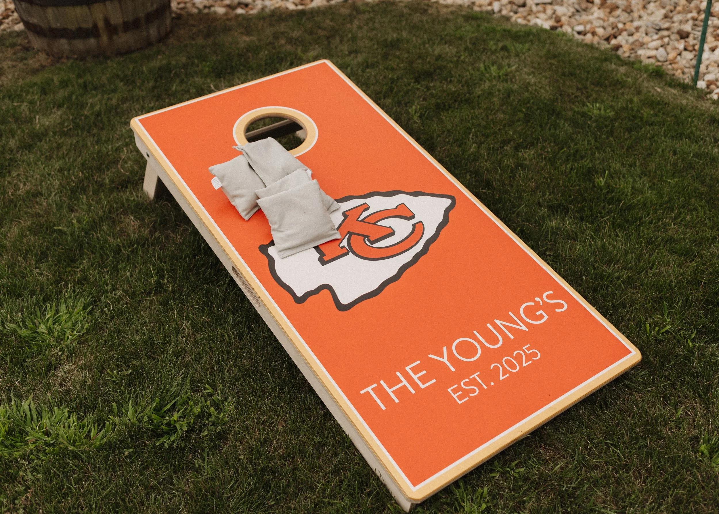 A cornhole game board on grass with the Kansas City Chiefs logo, white text reading "The Young's" and "Est. 2025," and three small white bags on top.