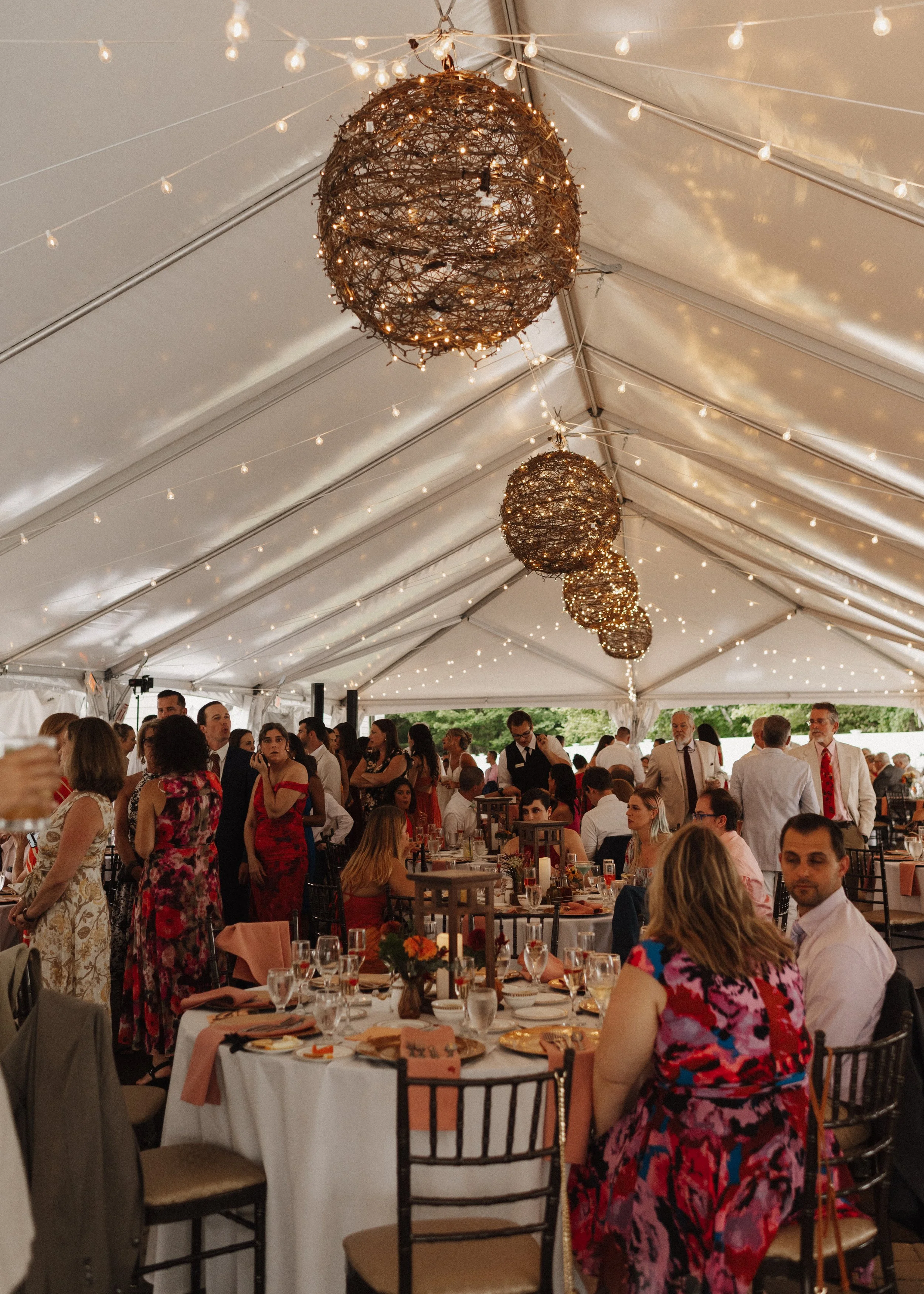 People gathered at a wedding reception inside a decorated tent with hanging string lights and large spherical twig chandeliers.