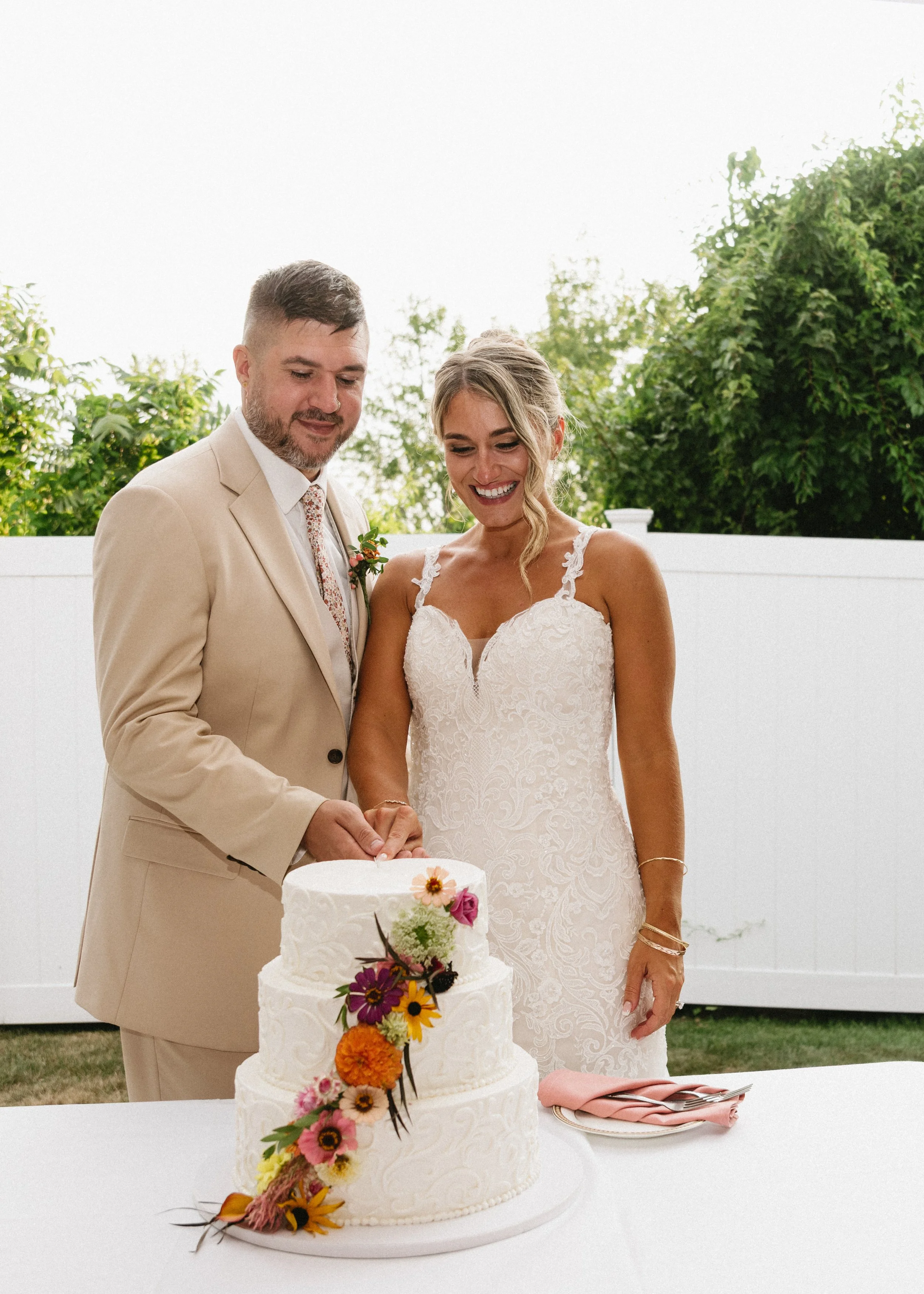 A couple in wedding attire cutting a three-tiered wedding cake decorated with colorful flowers, outdoors with greenery in the background.