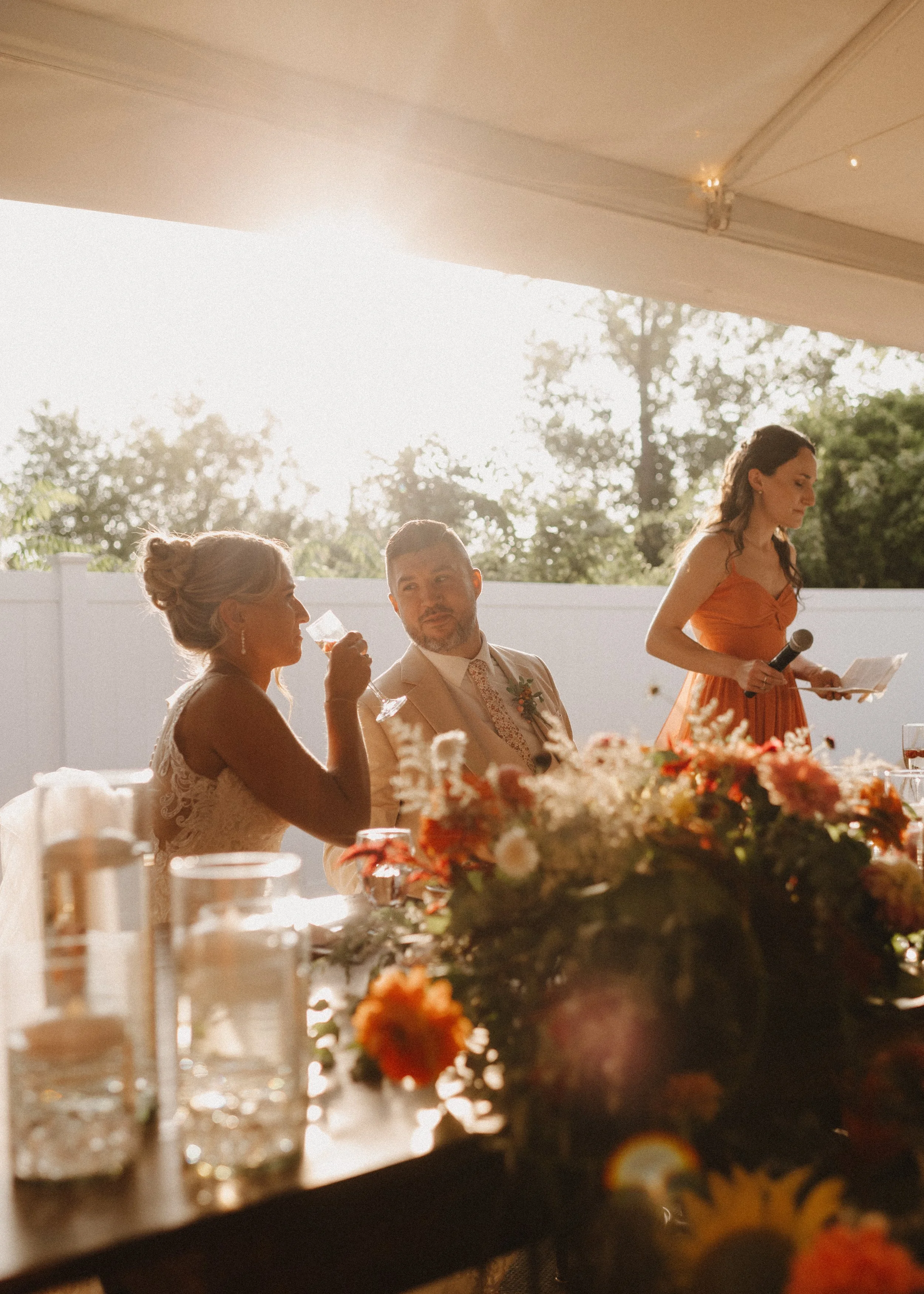A bride with a blonde updo and a groom with a beard at a wedding reception, sitting at a table with a large floral centerpiece, while a woman in an orange dress holds a microphone and reads from a paper under a canopy with sunlight in the background.