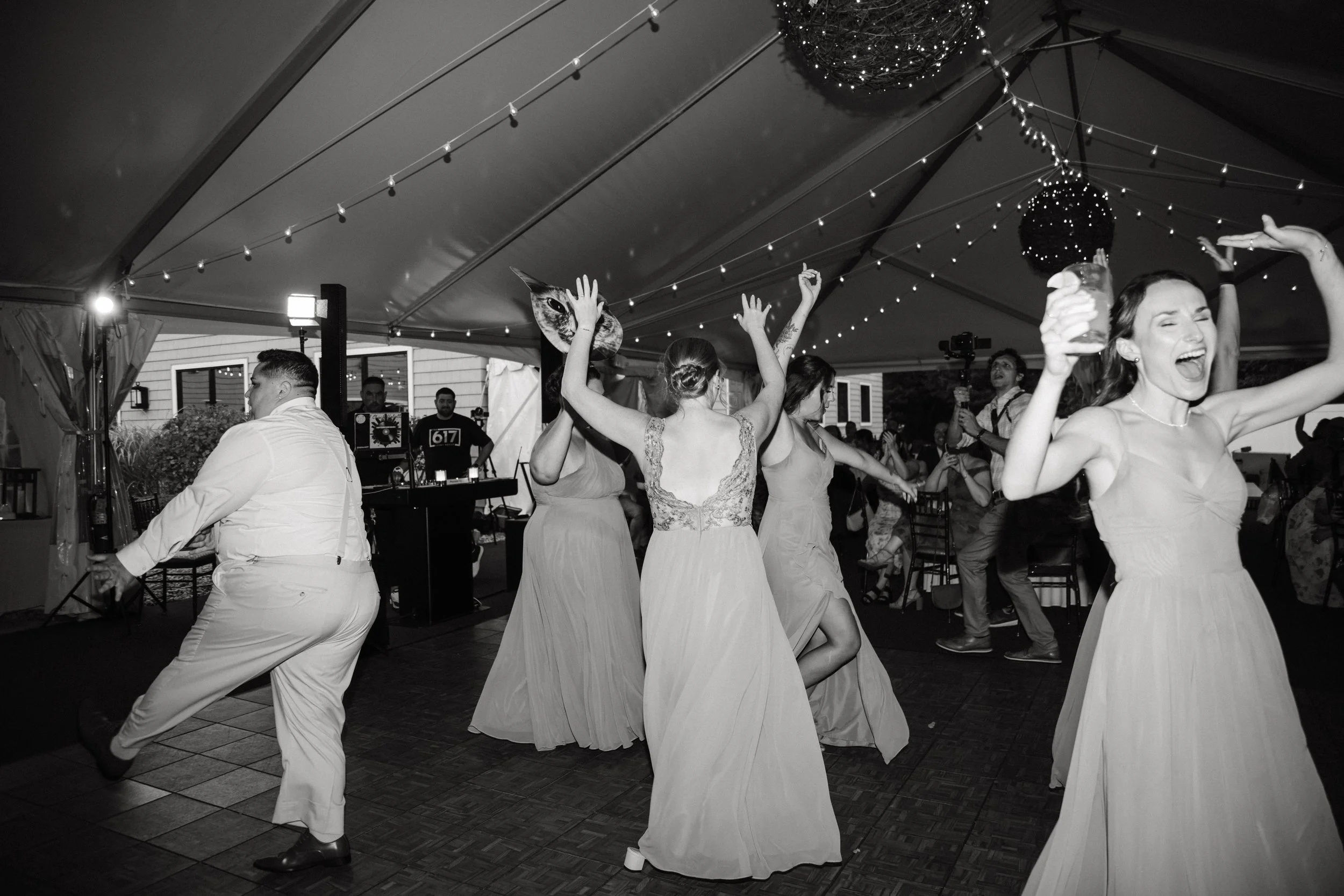People dancing and celebrating under a tent decorated with string lights at a wedding reception, with a DJ booth in the background.