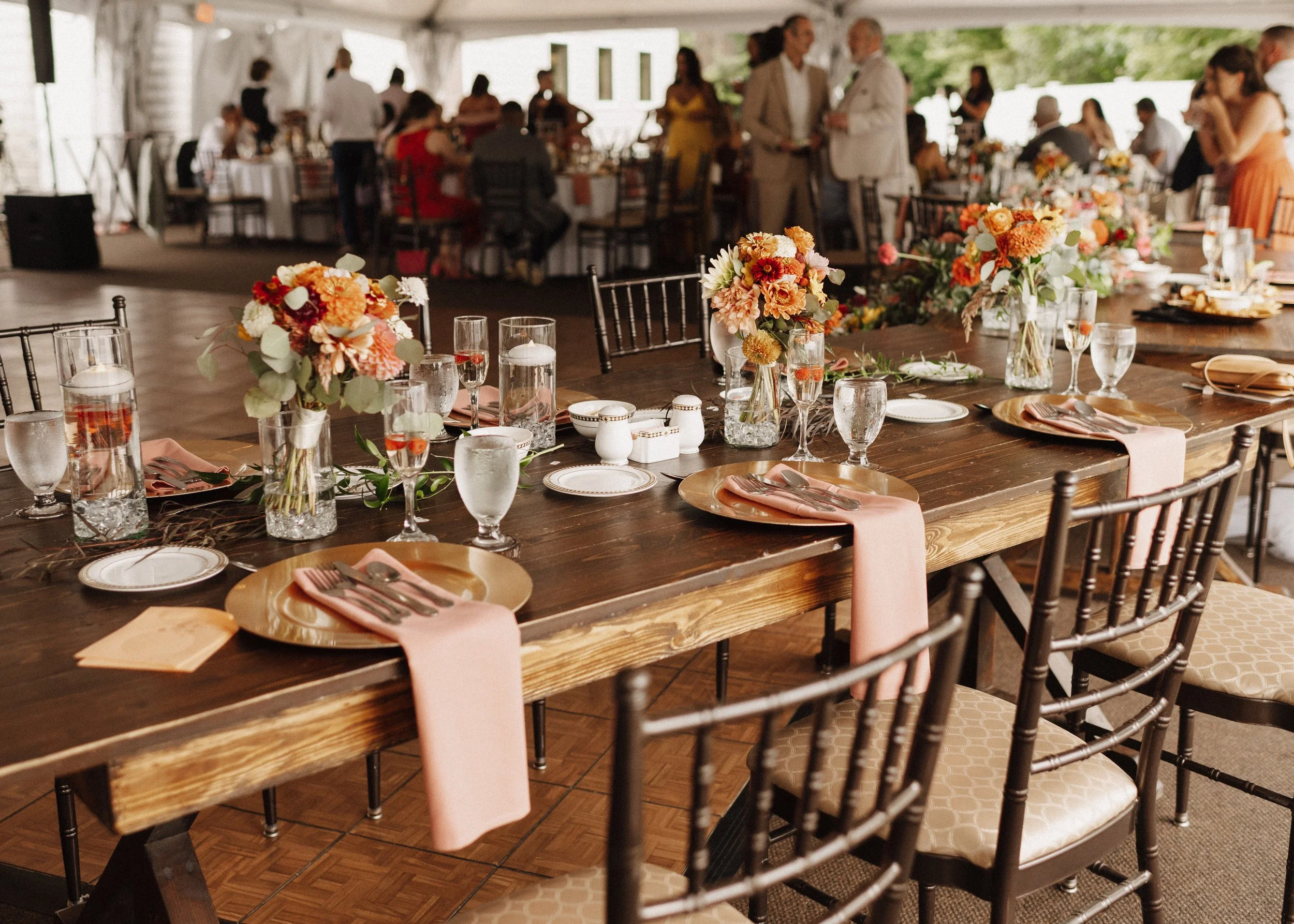 A decorated banquet table at an event, with floral centerpieces, plates, glasses, napkins, and silverware, set for a formal meal. In the background, people are mingling and seated at other tables under a large tent or open pavilion.