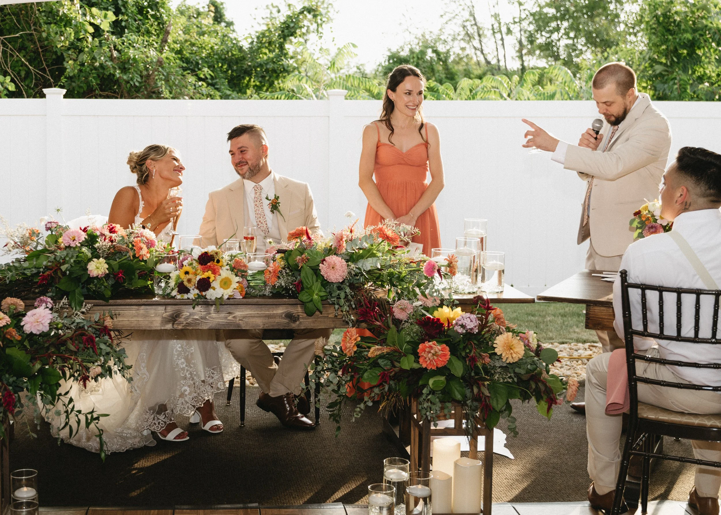 A wedding reception with a bride and groom sitting at a table decorated with flowers, while a woman in an orange dress stands speaking and another man in a beige suit makes a gesture with a microphone. Several guests are seated and the scene is outdo