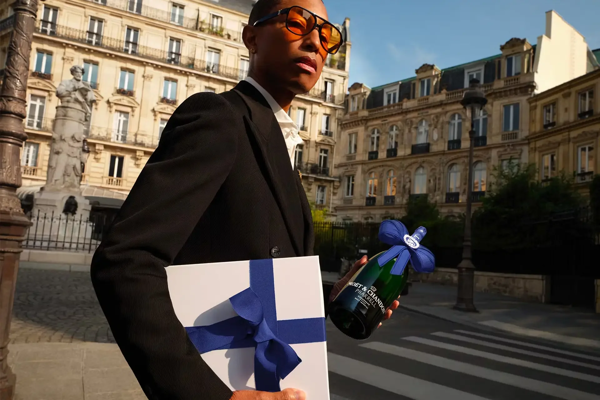Woman in a black blazer wearing orange sunglasses holding a bottle of Moët & Chandon champagne, a white and blue gift box, and a document, standing on a city street with historic Paris architecture in the background.