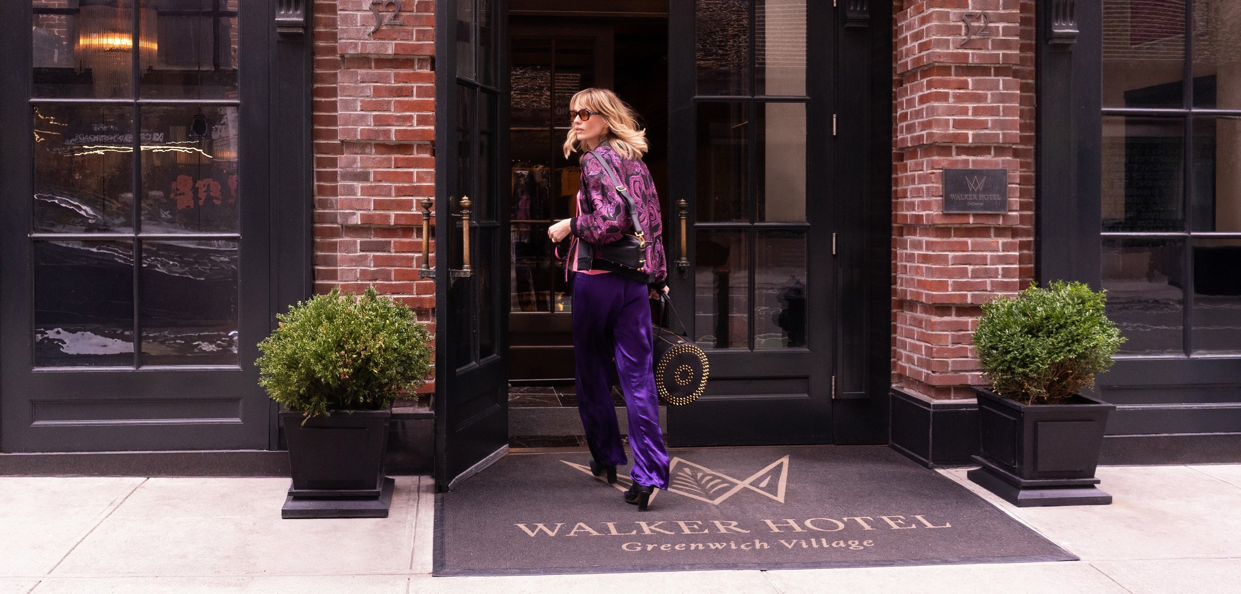 A woman with blonde hair wearing sunglasses, a purple patterned jacket, and purple satin pants is standing at the entrance of the Walker Hotel in Greenwich Village. She is holding a black handbag and balancing on high heels, next to potted bushes and black-framed windows and doors.