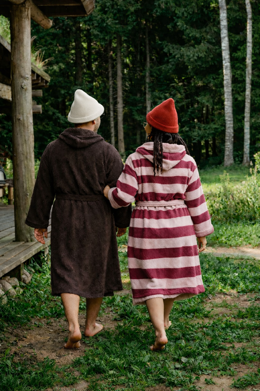 A man and woman walking barefoot on a garden path, dressed in cozy robes and hats, surrounded by green trees and plants.