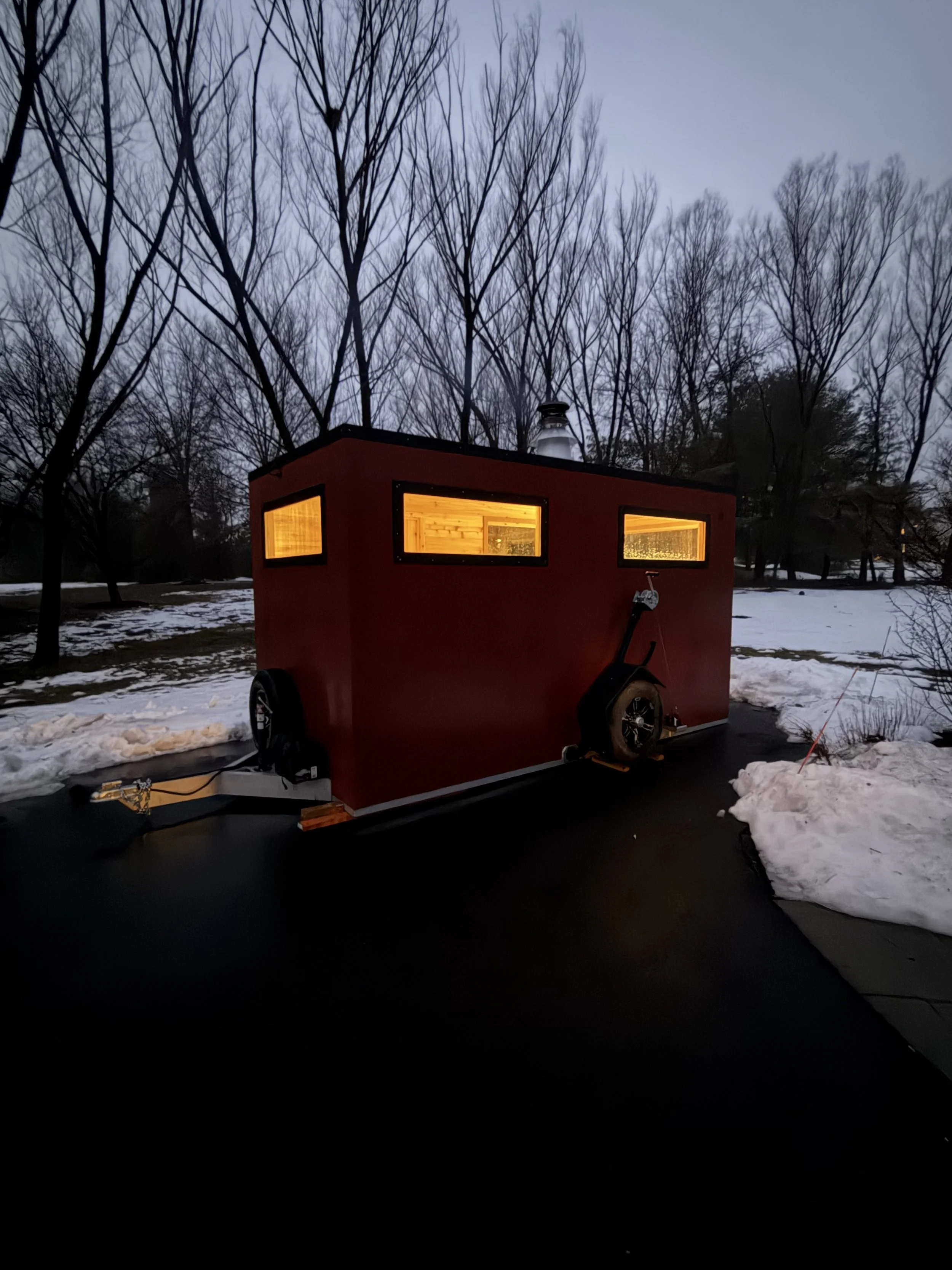 A small red camper trailer with illuminated interior windows parked on a dark asphalt driveway, surrounded by snow and leafless trees on a cloudy evening.