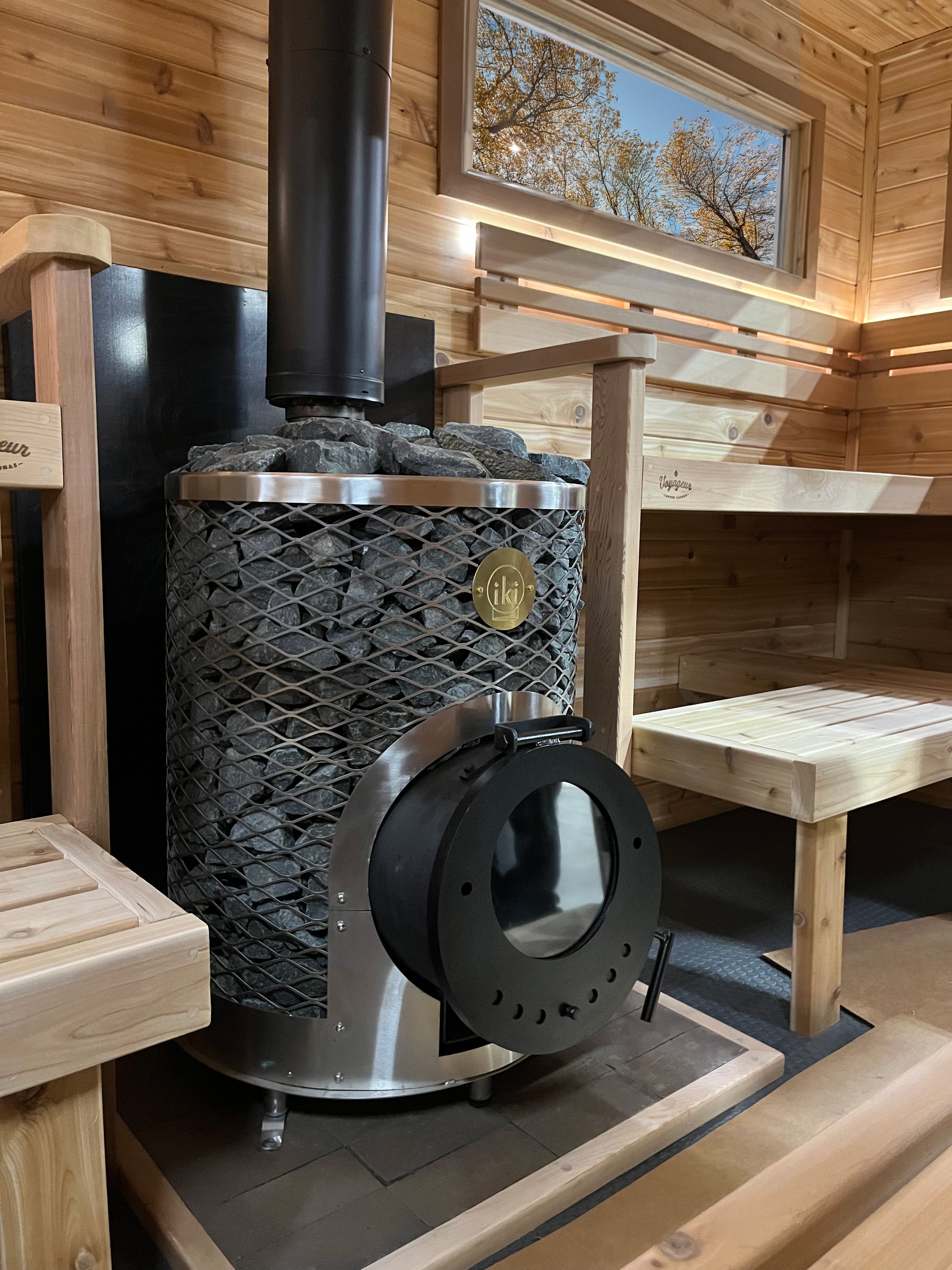 Inside a wooden sauna room with a wood stove filled with stones, a black pipe, a window showing trees outside, and wooden benches.