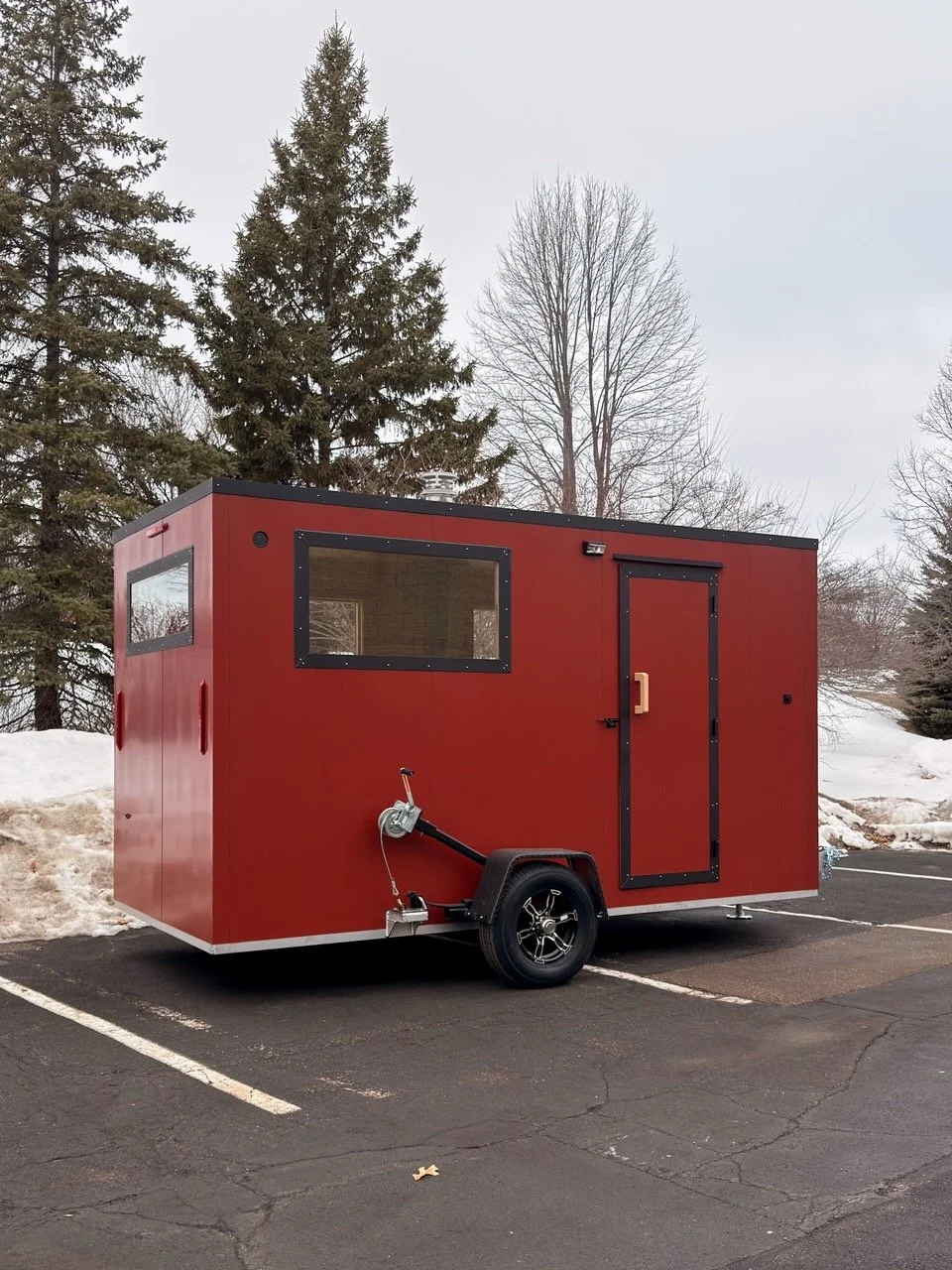 Red food truck trailer parked in an empty parking lot with snow piles and trees in the background.
