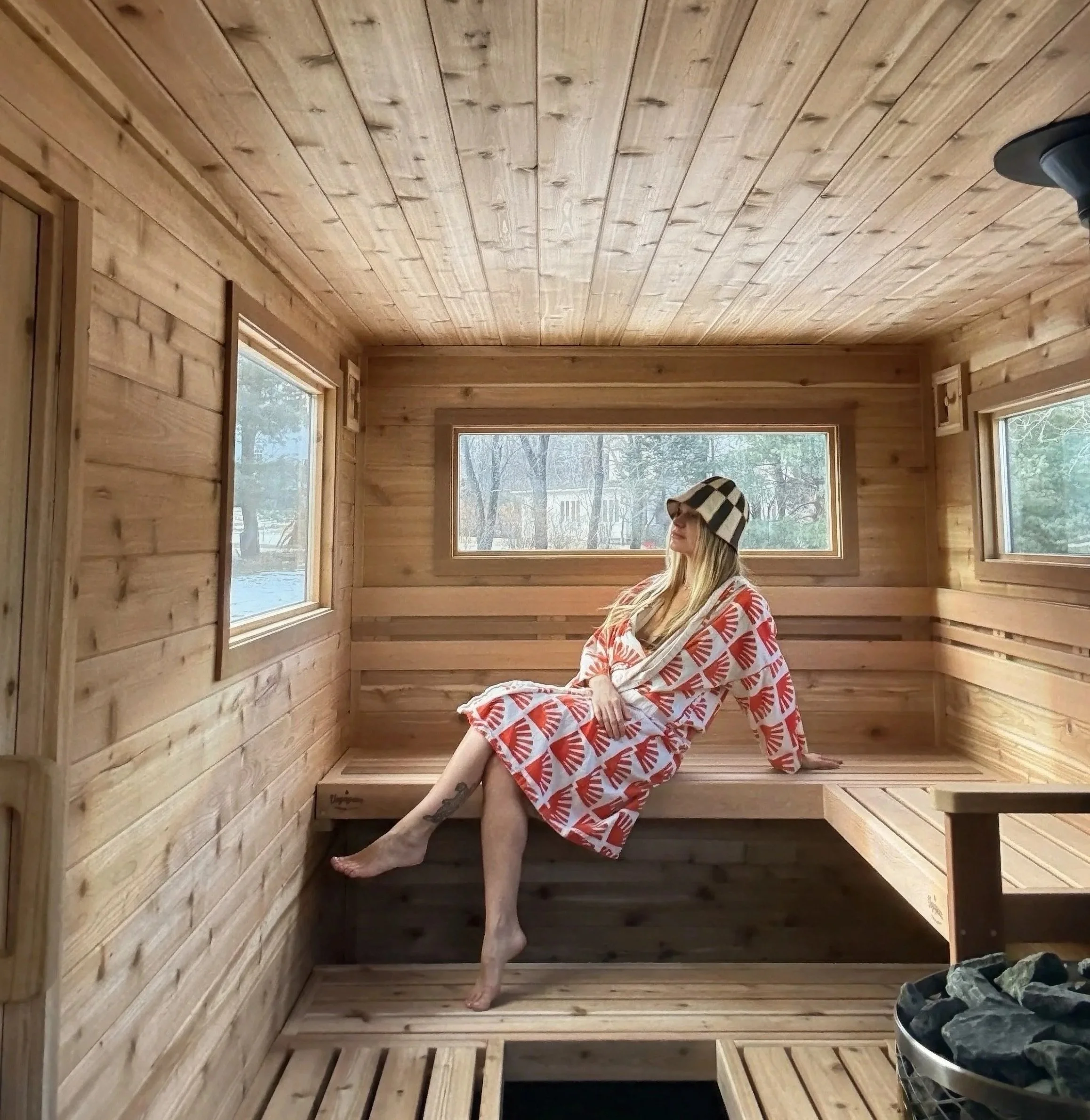 A woman sitting in a wood-paneled sauna, wearing a patterned towel and a checkered hat, with her legs crossed and one hand resting on the bench, looking outside through the windows.