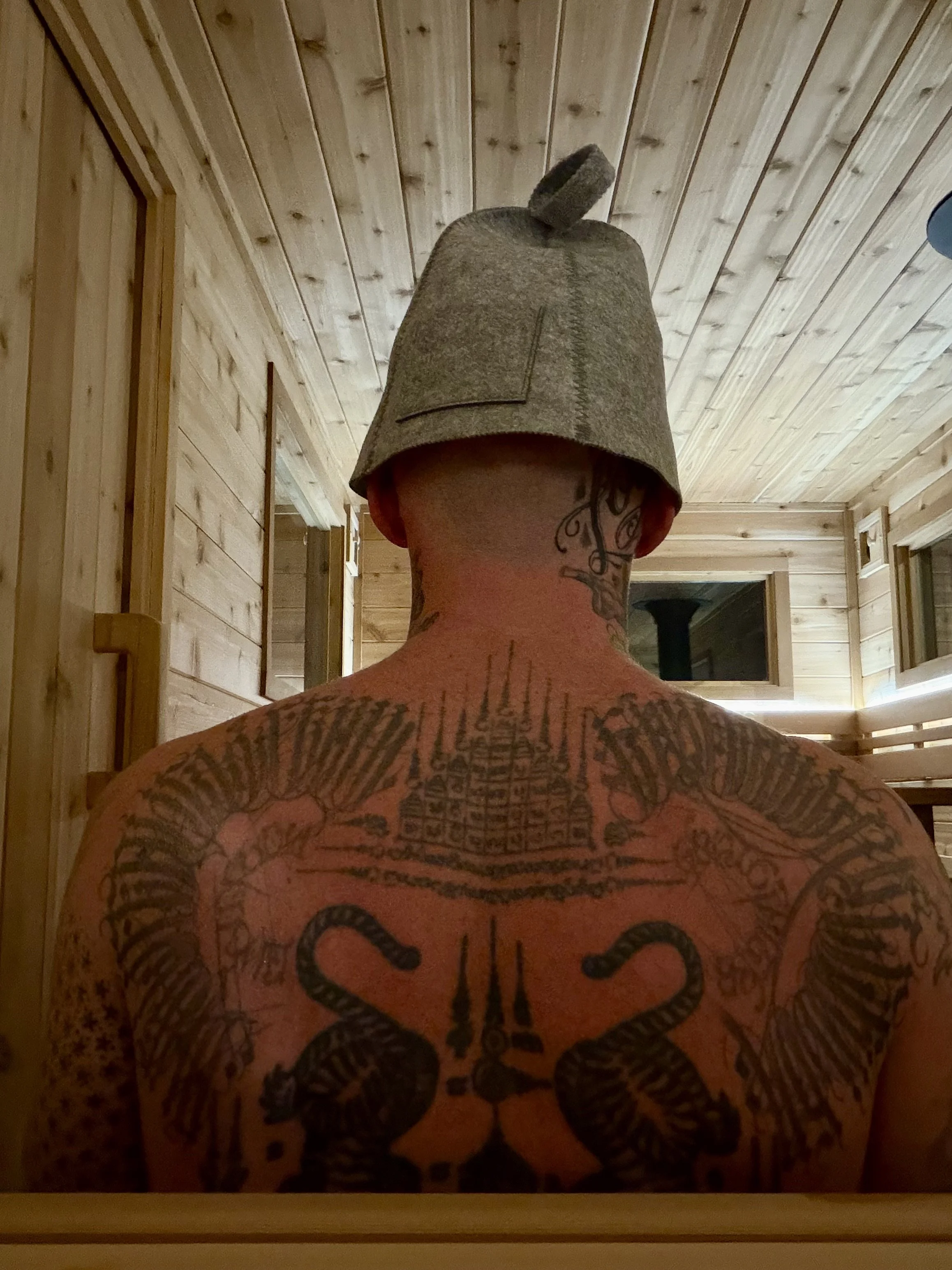 Back of a man with tattoos, wearing a gray sleeping cap, sitting in a wooden room with a wood-paneled ceiling and walls.