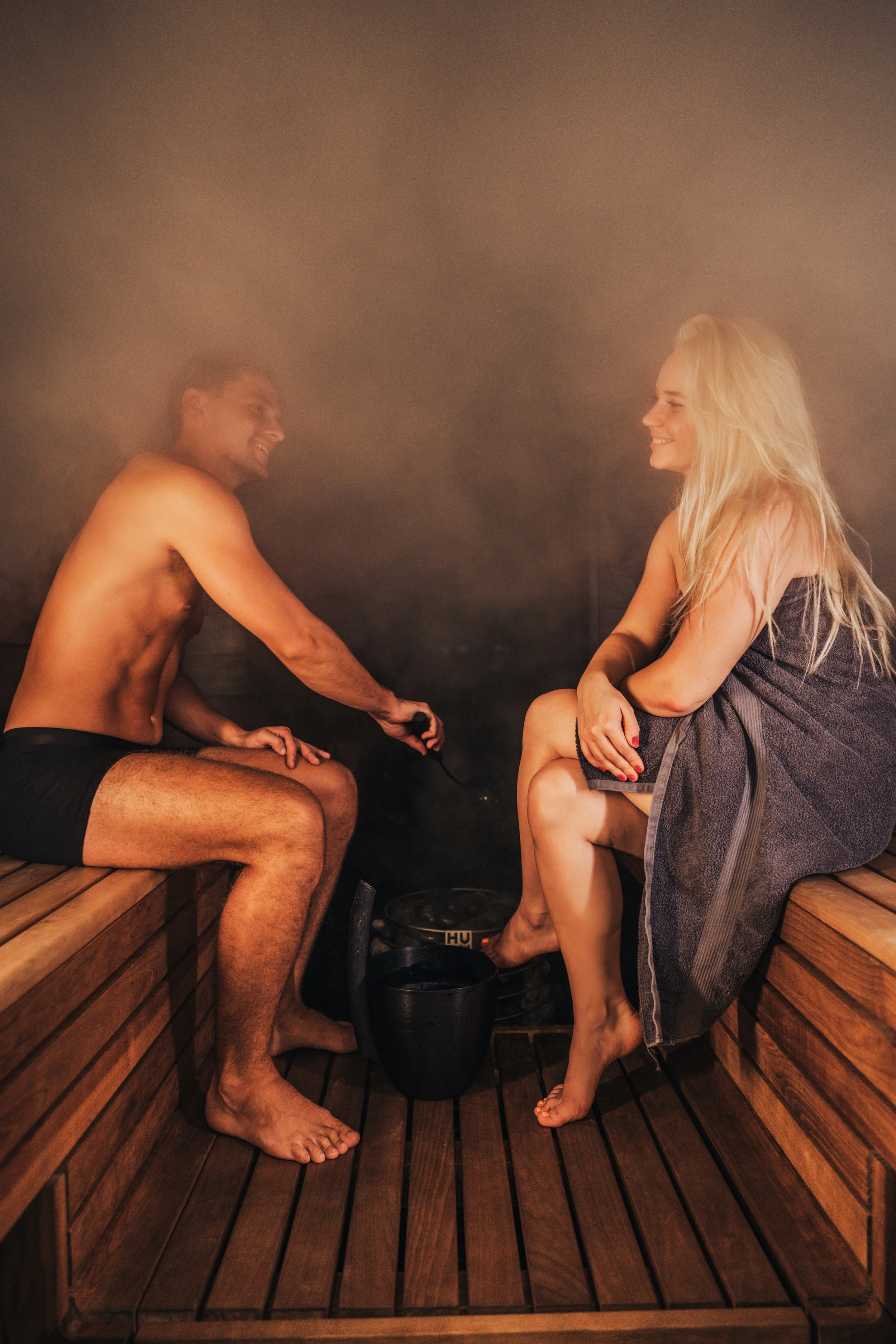 A man and a woman sitting in a sauna, smiling at each other. The man is using a handheld device, and both are dressed in towels.