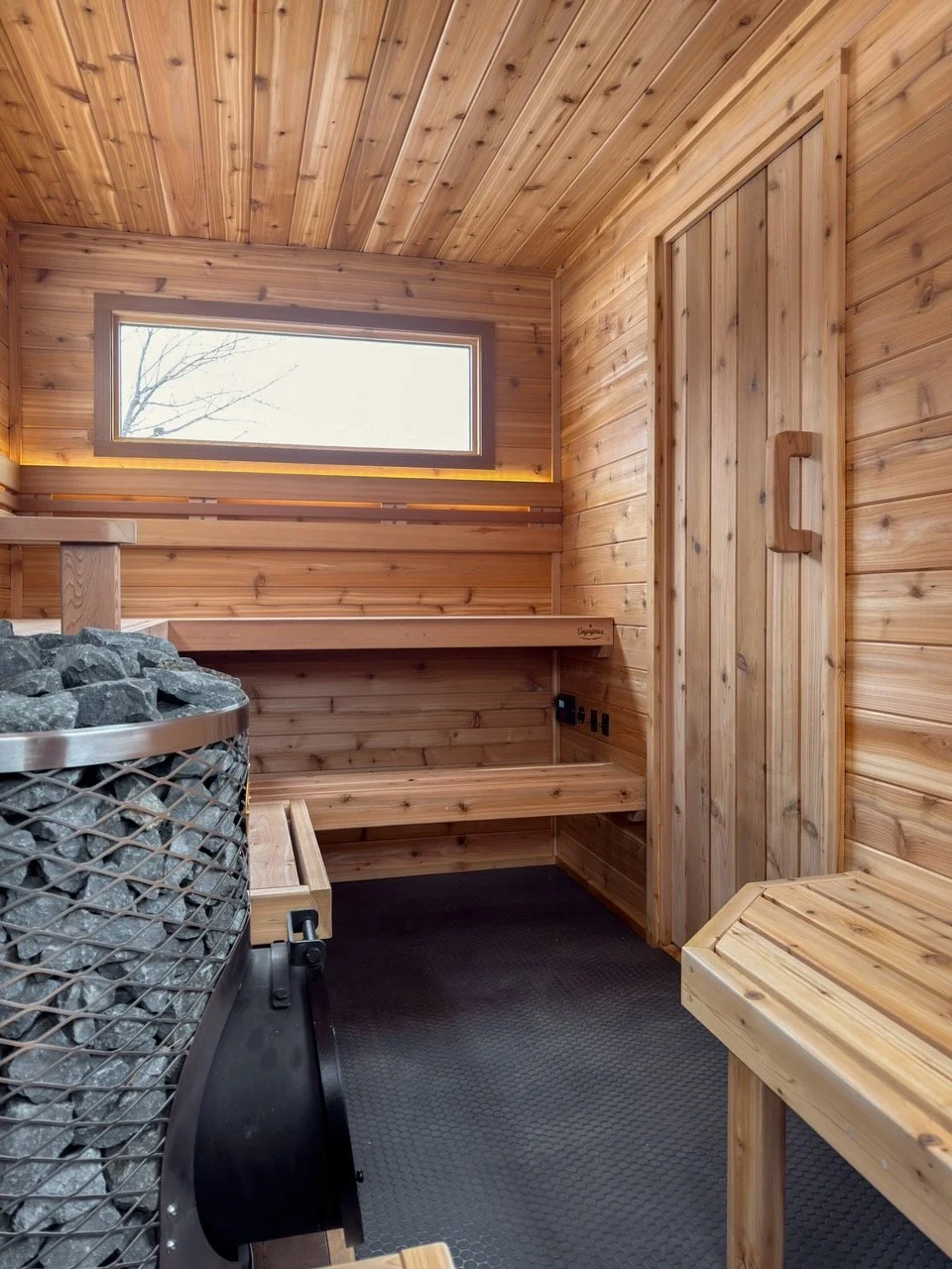 Window view of a wooden sauna interior with benches, a heater with stones, and a small table.
