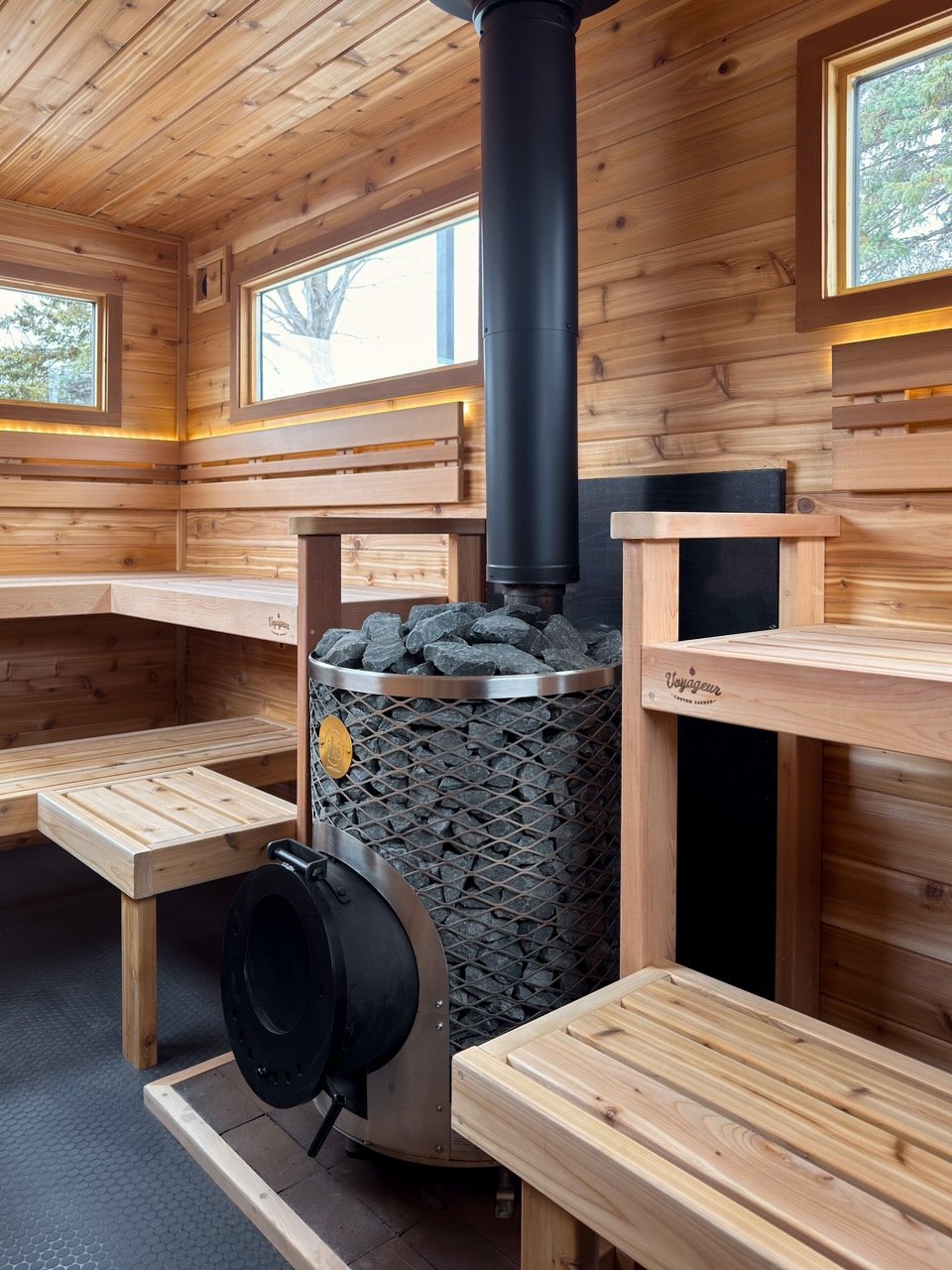 Interior of a wooden mobile sauna trailer with benches, a wood stove filled with rocks, and three small windows.