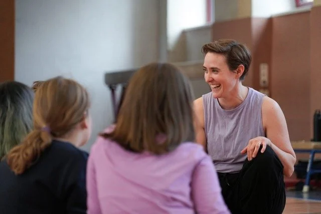Gemma Braun, Kids-Trainerin im Mini Movers Club Berlin, sitzt lächelnd auf dem Boden eines hellen Sportstudios für Kinderkurse.