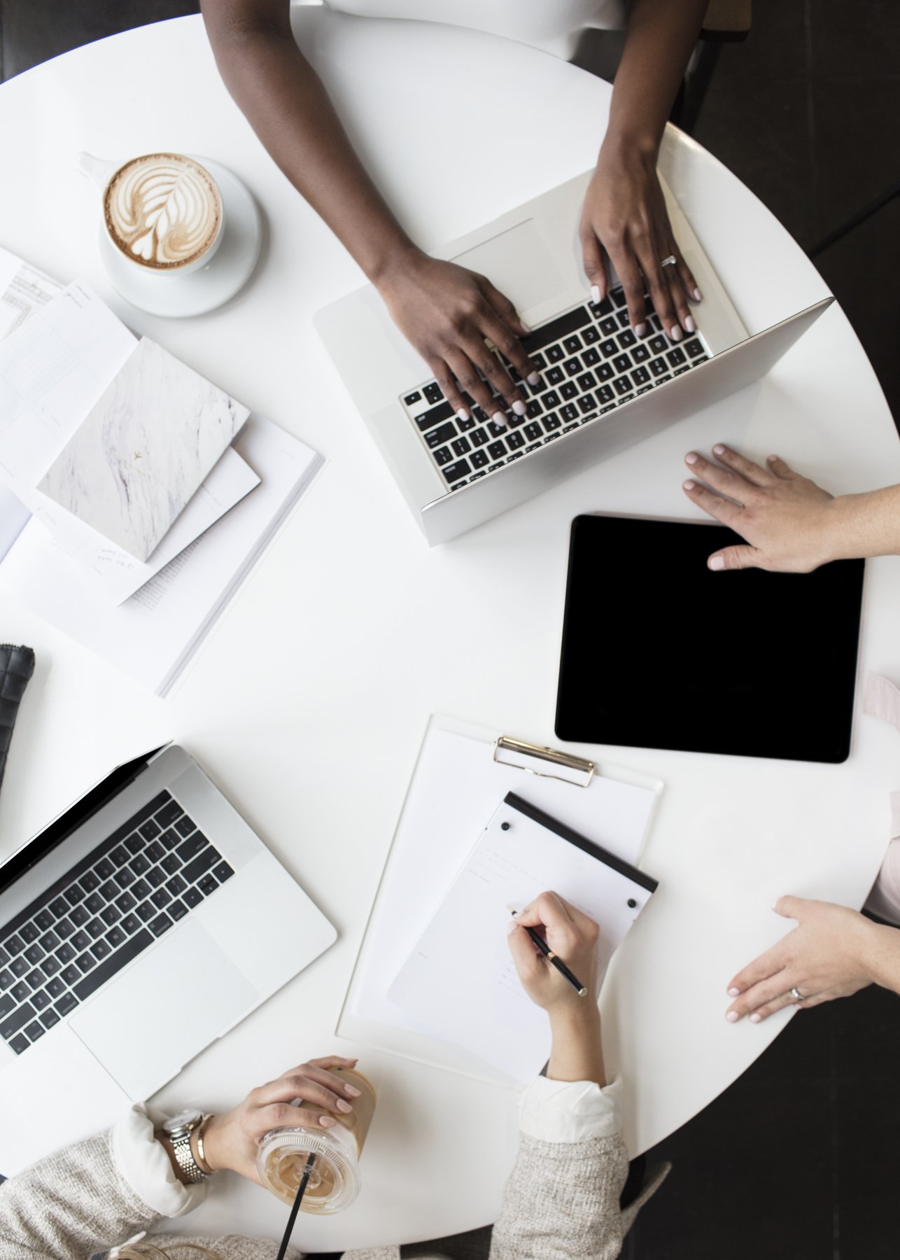 Overhead view of three people working at a white round table with laptops, a tablet, notebooks, documents, a coffee cup, and a hand holding a beverage cup with a straw.