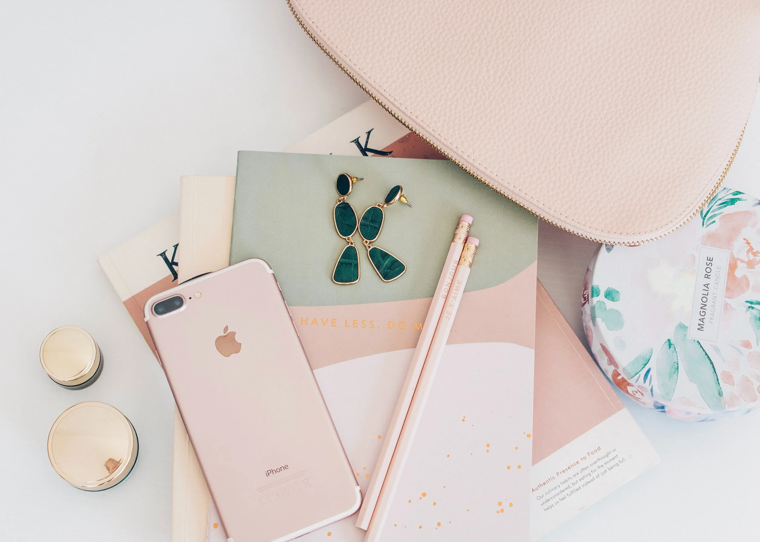 Flat lay of a pink iPhone, gold-rimmed earrings with green stones, pink pencils, a pink textured purse, a floral scented candle, and magazines on a white surface.