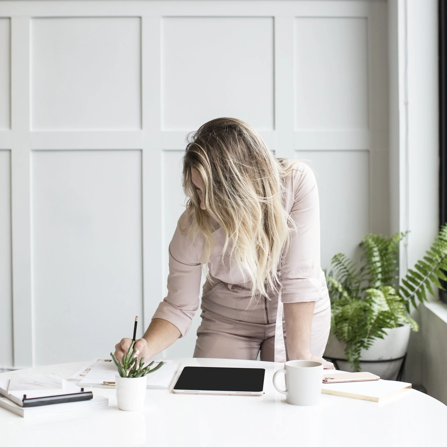 Woman with long blonde hair leaning over a white desk, writing in a notebook, with a tablet, mugs, and plants around her.