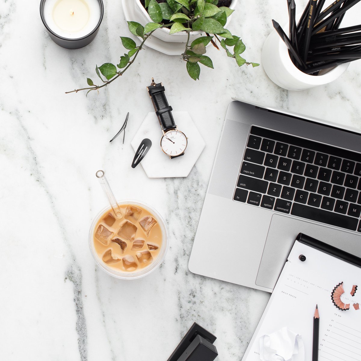 A white marble desk with a laptop, iced coffee, watch, notebook, pens, pencil sharpener, plant, candle, hairpins, and a hair clip.