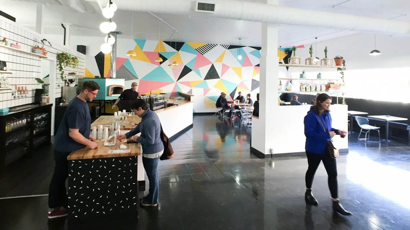 Interior of a modern cafe featuring a colorful geometric mural on the wall, a counter with baristas, and customers seated at tables.