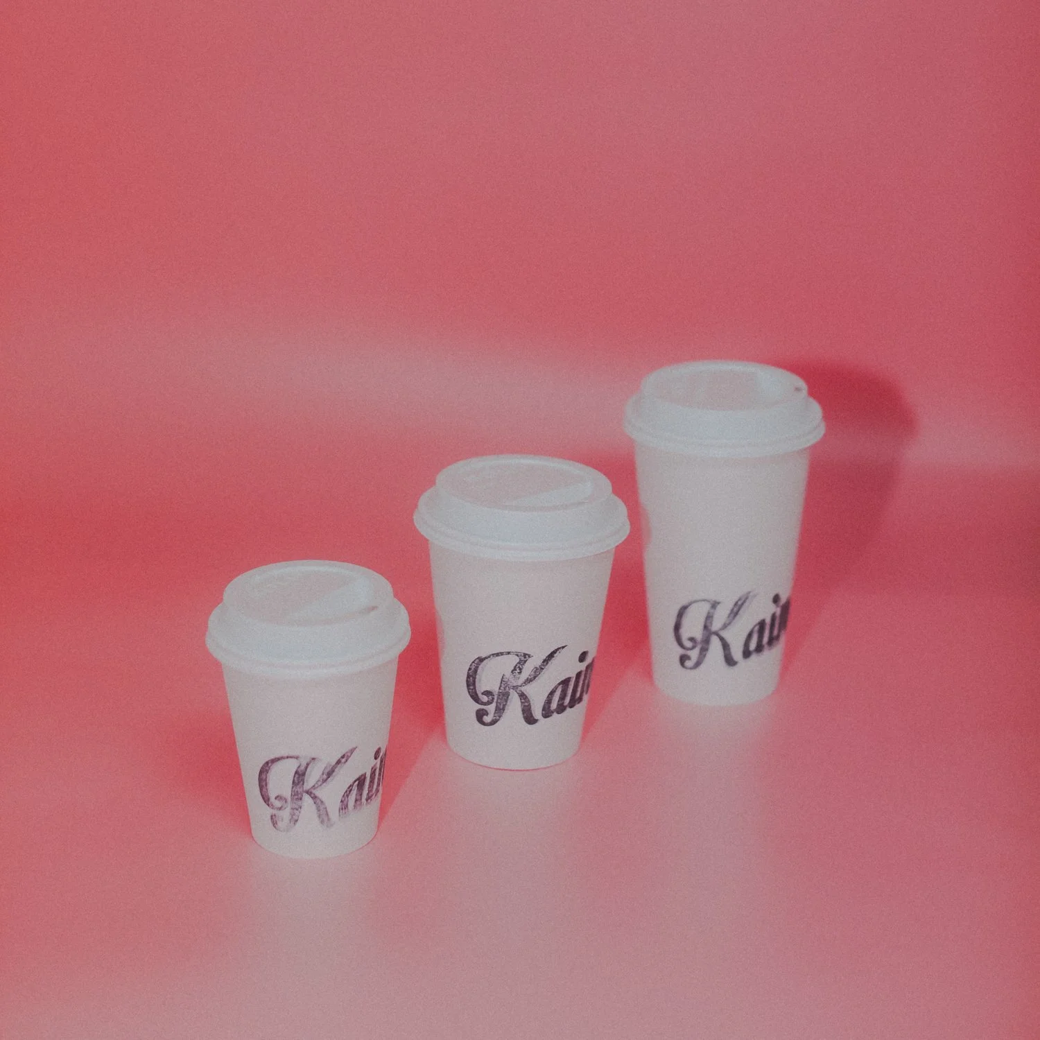 Three white disposable coffee cups with white lids, each labeled with the word 'Kai' in black cursive, set against a pink background.