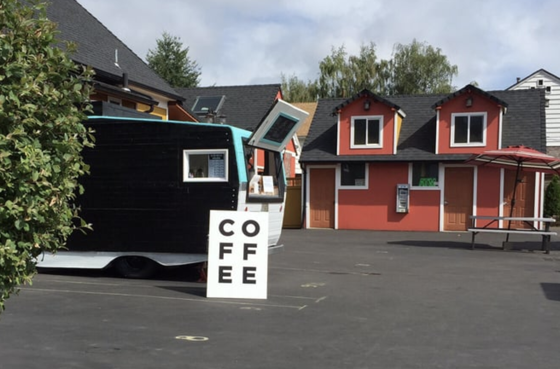 A small coffee stand shaped like a boat with a sign reading 'COFFEE' in front of a colorful building, with an umbrella and picnic table nearby.