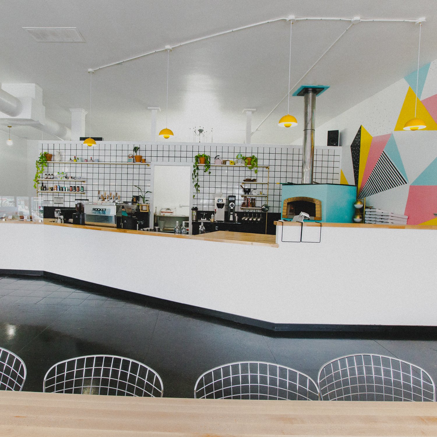 Modern cafe with a white tiled wall, colorful geometric mural, yellow pendant lights, and a pizza oven.