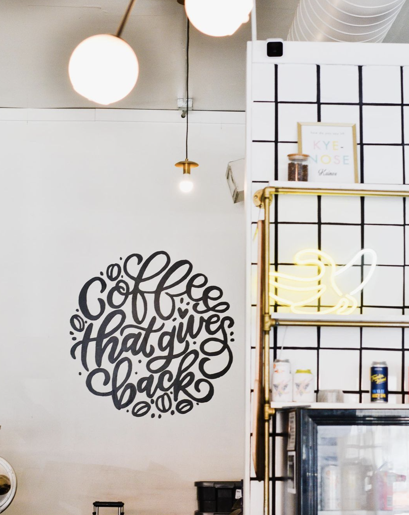 Interior of a coffee shop with a white wall featuring a black mural that says 'coffee that gives back' in stylized lettering. There are hanging light fixtures and a neon sign of a coffee cup on a black grid wall.