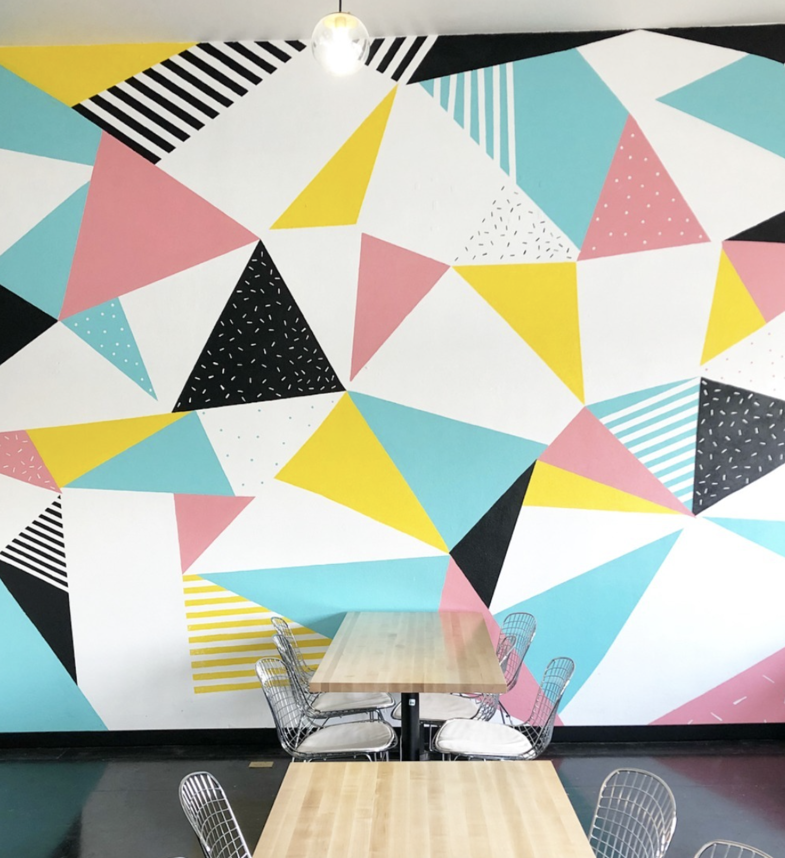 Colorful geometric mural with triangles in pink, yellow, black, white, and turquoise, with various patterns, in a modern cafe or dining area with wooden tables and wireframe chairs.
